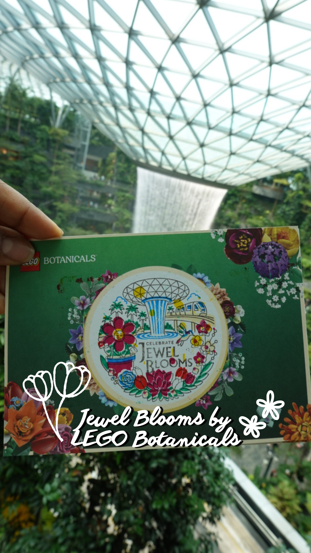 A person holding a Jewel Blooms by LEGO Botanicals booklet in front of a lush indoor garden with a large glass roof and a waterfall.