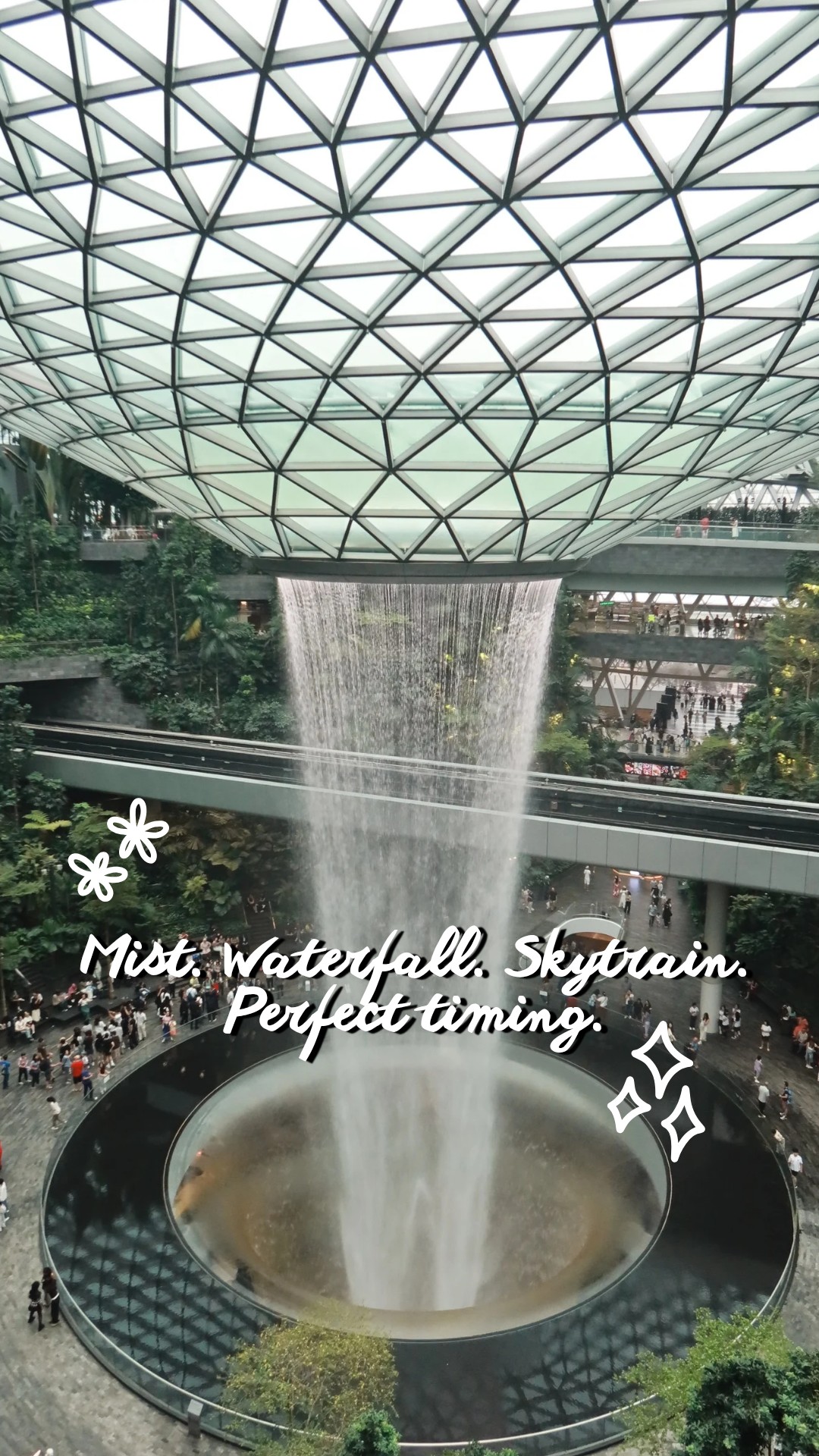 Interior view of the Jewel Changi Airport in Singapore featuring the Rain Vortex, the world's tallest indoor waterfall, with a glass and steel skylight above and a mist effect around the waterfall, surrounded by lush greenery and visitors.