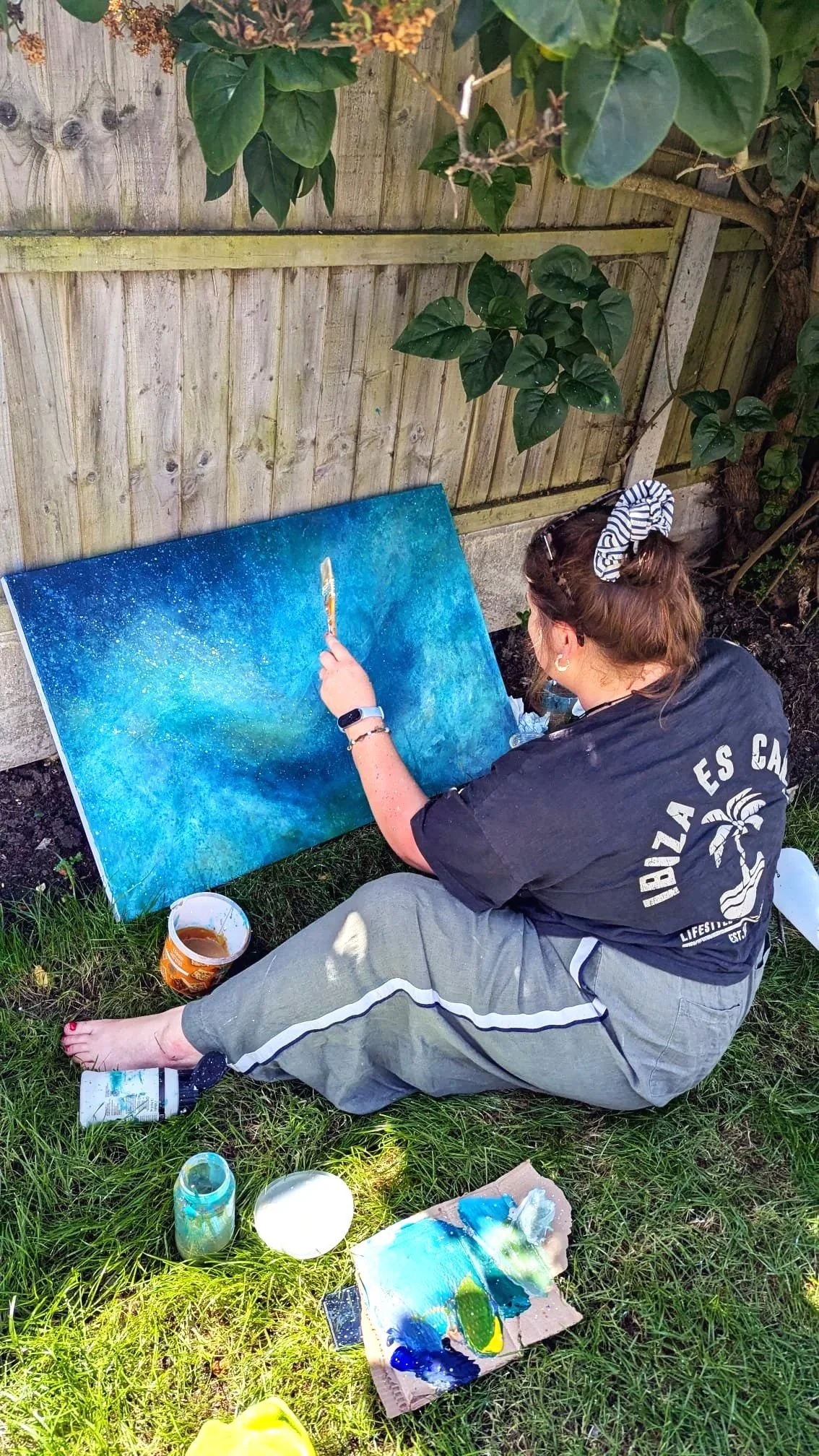 Artist Alice Chapman in her garden painting a vibrant seascape. Painting supplies, including a palette with blue and green paint, jars, and cups, are around her.