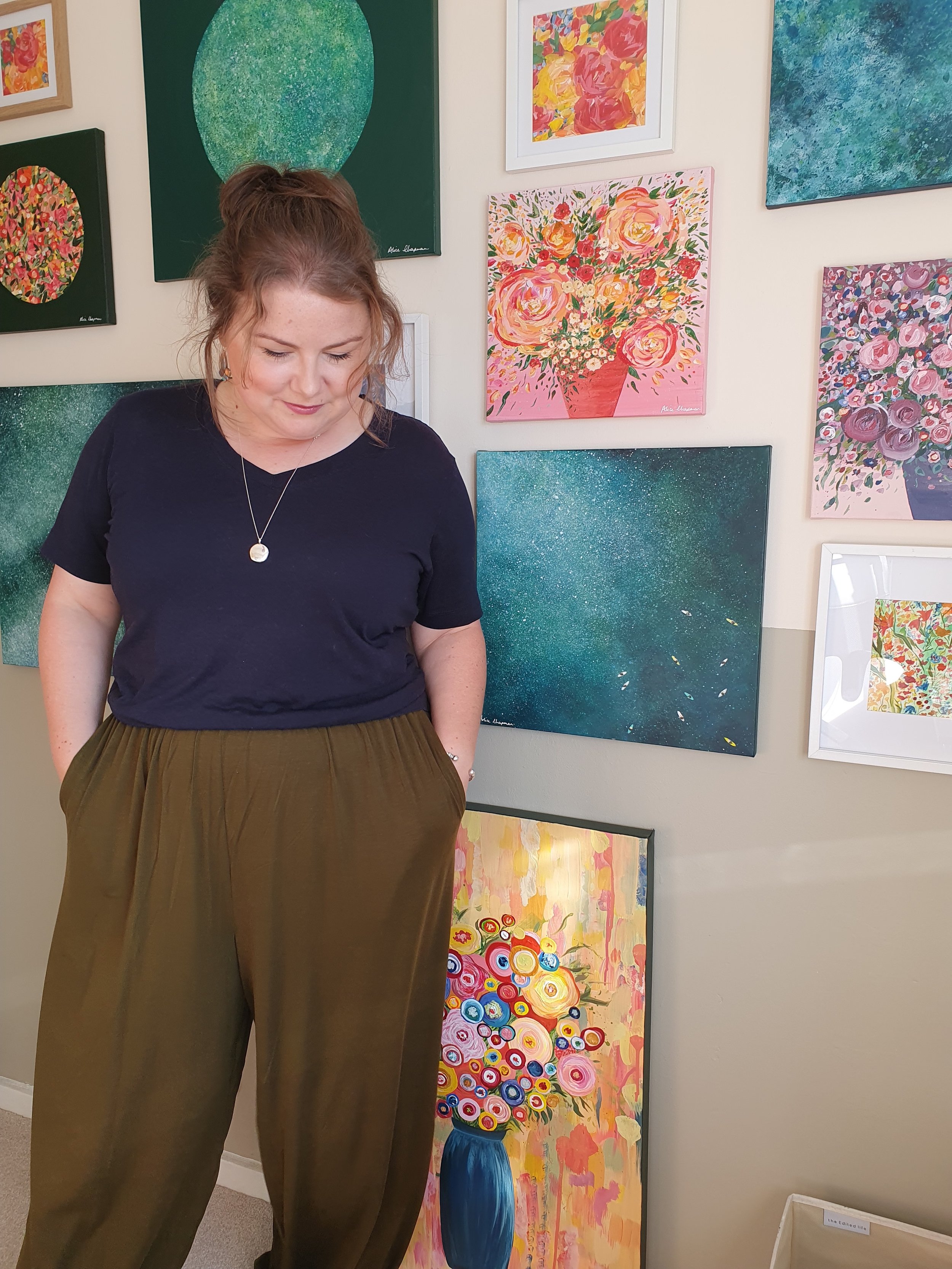 Artist, Alice Chapman stands in front of a wall decorated with colourful floral and seascape paintings in her studio in Cheltenham.
