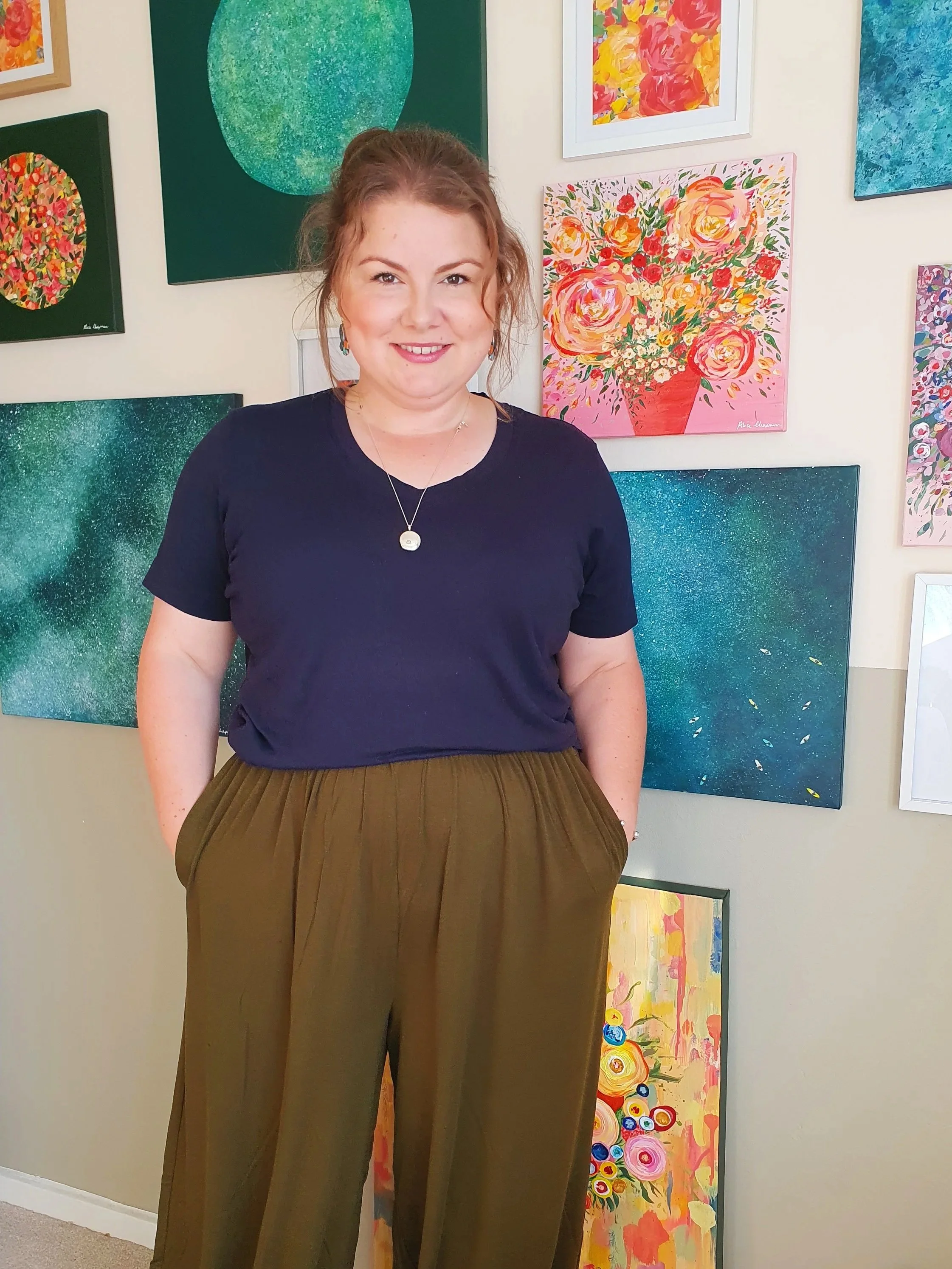 Artist, Alice Chapman is standing in front of her studio wall in her home in Gloucestershire. The wall is decorated by vibrant and mesmerising original paintings.