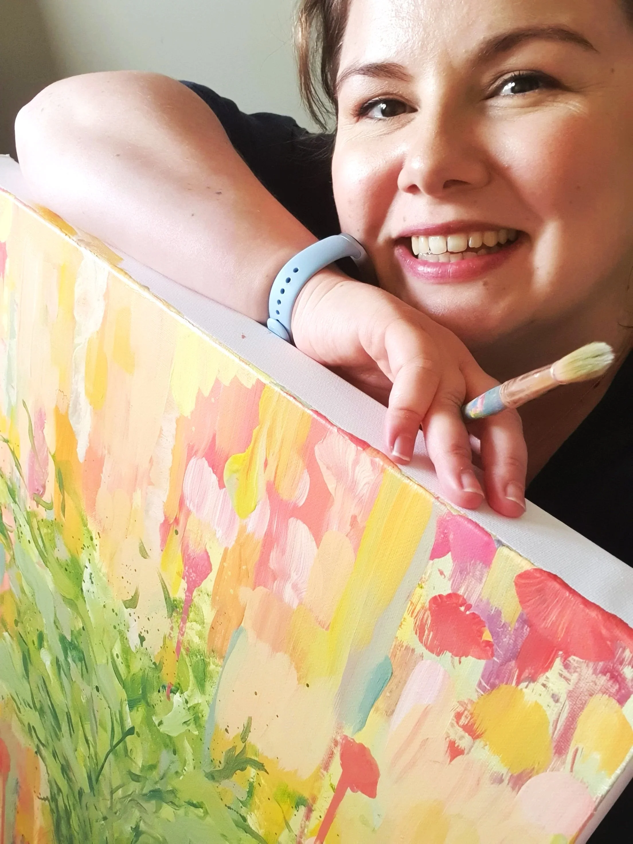 Artist Alice Chapman smiling and resting her head on her arm, which is on one of her colourful, abstract paintings. It is unfinished but capturing the joy she experiences when in her studio.