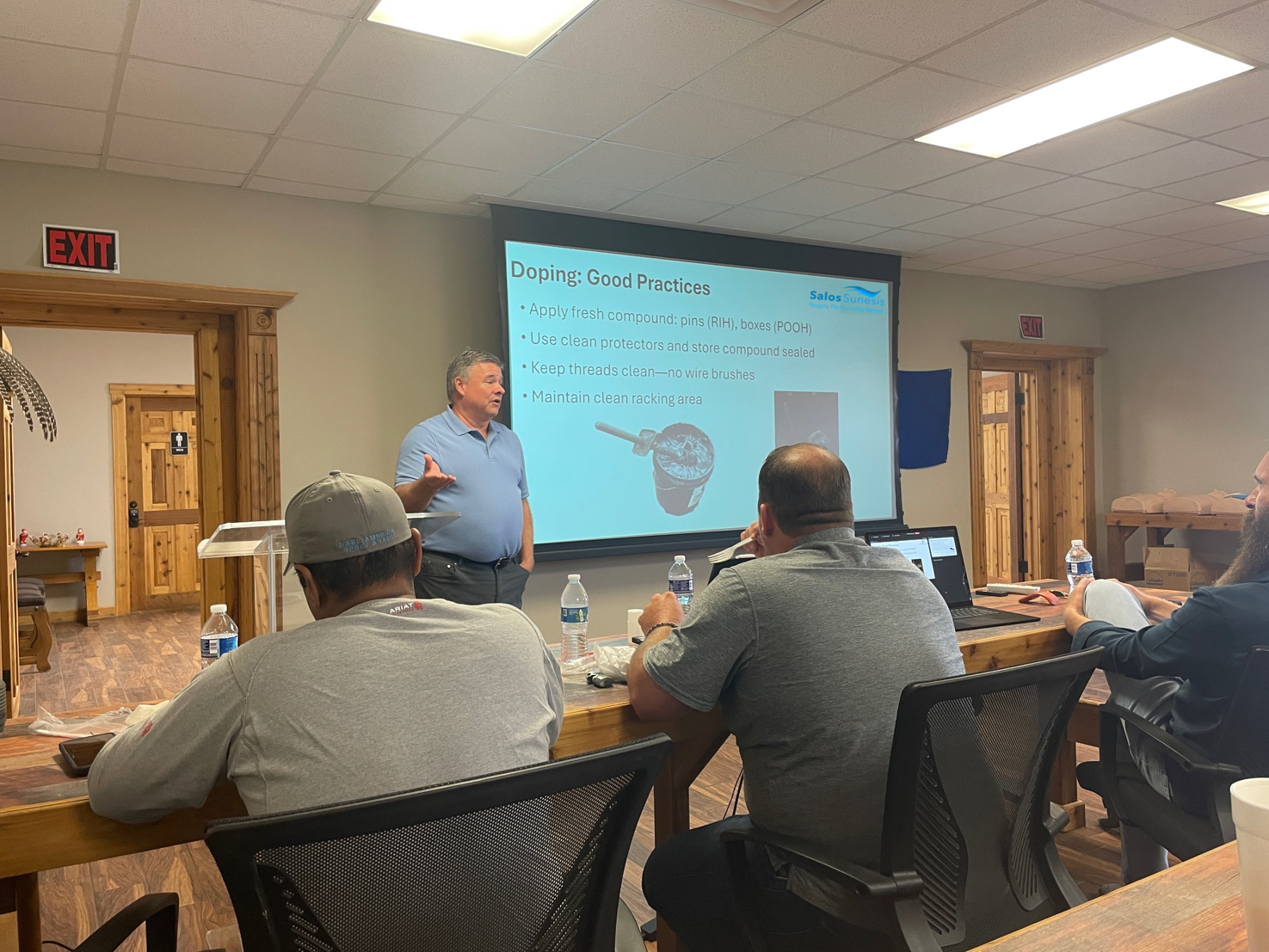 A man in a blue polo shirt is giving a presentation in a conference room with several attendees seated at a wooden table, with a slide on doping good practices projected behind him.