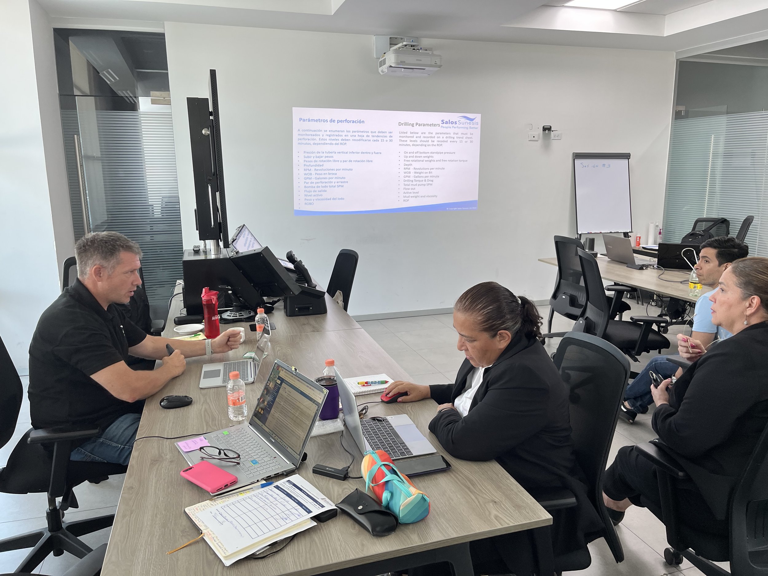 People sitting around a conference table in a meeting room, engaged in a discussion. A large screen on the wall displays a presentation slide titled 'Parámetros de perforación' with English translation, and several people have laptops and notebooks in front of them.