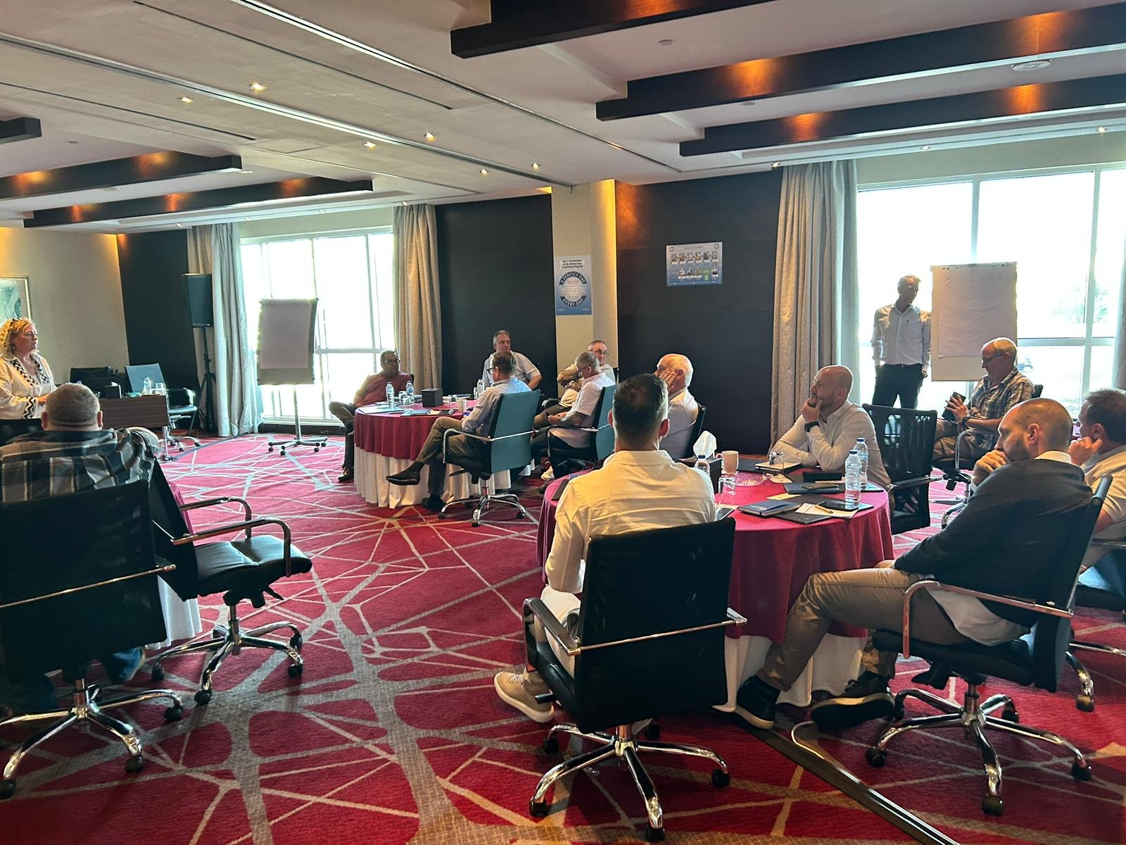 Business meeting or conference in a high-rise hotel with participants seated at round tables, whiteboard, and presentation in a bright, modern conference room.