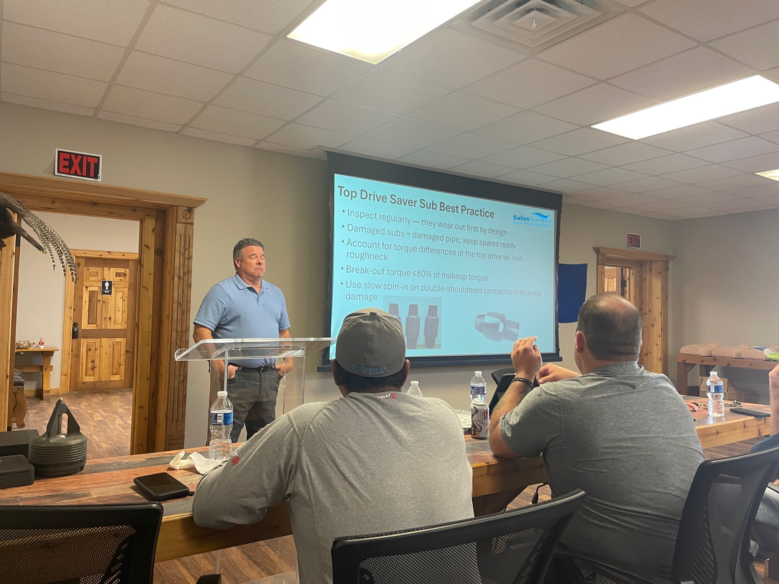 A man in a light blue polo shirt giving a presentation in a conference room. The presentation slide on the screen displays tips about top drive saver sub best practices. Several audience members are seated at a wooden table with water bottles and electronic devices.