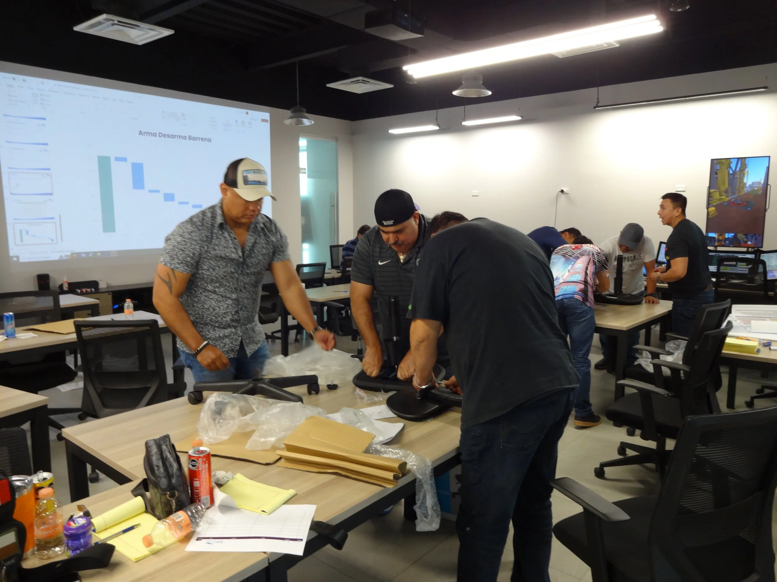 Group of people working at tables in a modern office or classroom setting, with a large screen displaying a presentation and a monitor airing a game in the background.