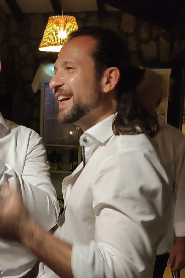 A smiling man with long hair and a beard, wearing a white shirt, in a warmly lit indoor setting.