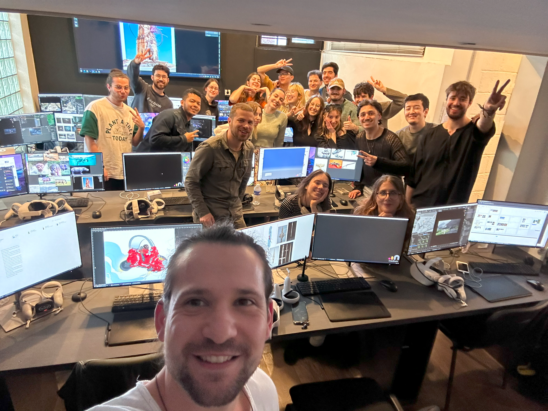Group of people gathered in an office with multiple computer monitors, some smiling and making peace signs, taking group photo.