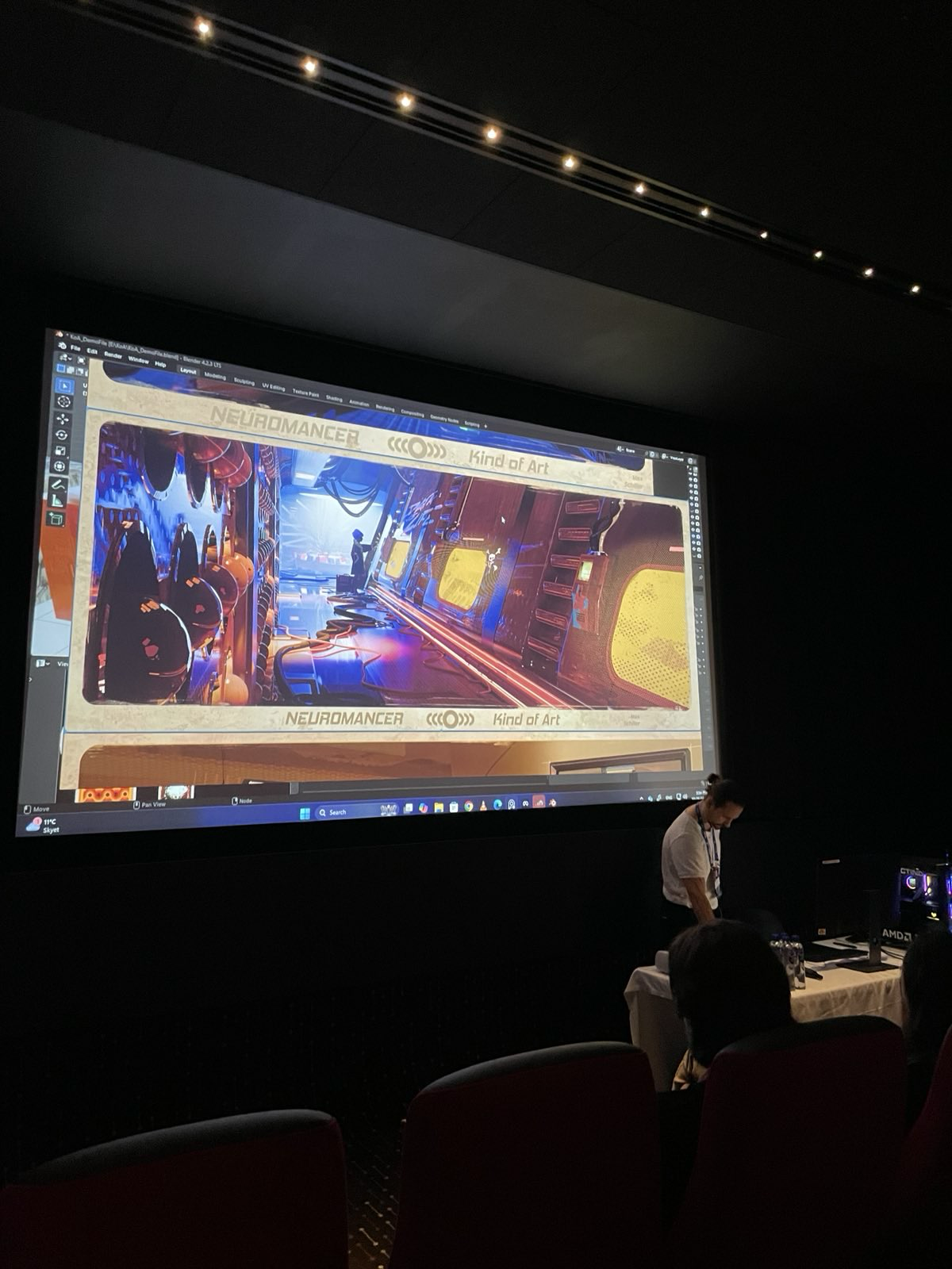 A presentation screen in a dark room displaying a digital illustration of a futuristic, neon-lit interior space with advanced technology and a person standing inside. A person is standing in front of the screen, and several audience members are seated and observing.