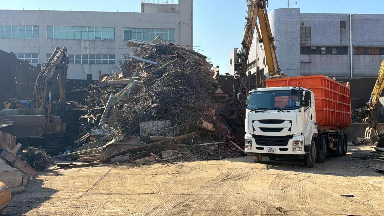 Construction site with excavators and a pile of scrap metal, with a white truck nearby, in front of industrial buildings.