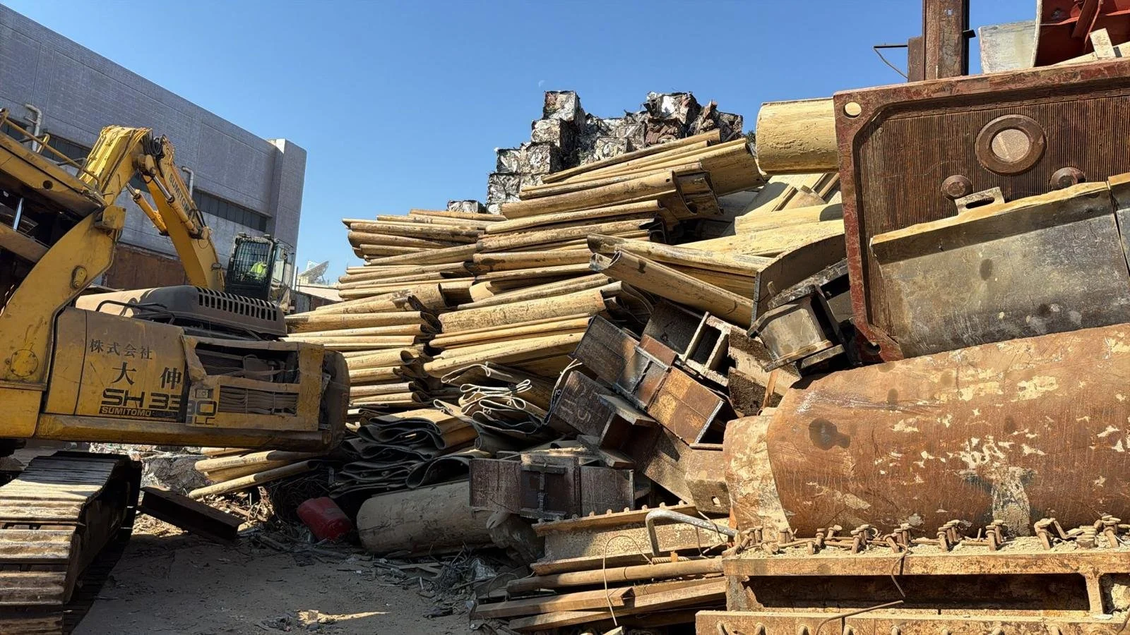Construction site with a yellow bulldozer, metal pipes, rusted machinery, and piled scrap metal under a clear blue sky.
