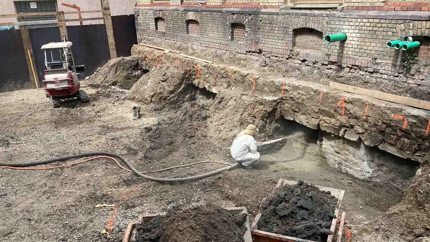 Bauarbeiter reinigt eine Grube mit einem Hochdruckreiniger, um die Wandbesonderheiten sichtbar zu machen. Das Baugelände ist mit Sand und Erde bedeckt, es gibt Rohre an der Wand. Eine Baufahrzeug steht im Hintergrund.