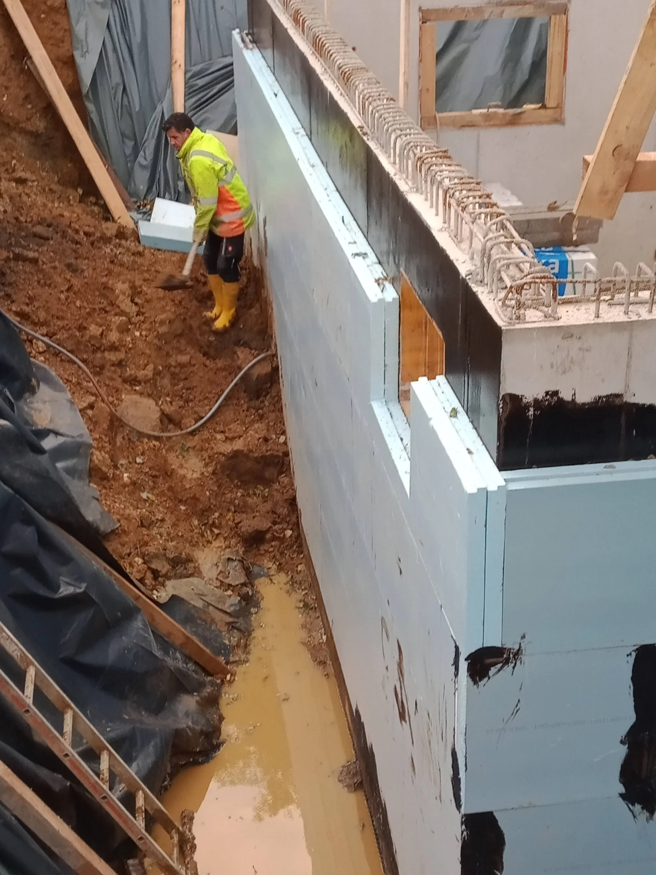 Ein Bauarbeiter mit gelber Warnjacke und Gummistiefeln arbeitet in einem Haus im Bau. Er schaufelt Erde in einem Graben neben einer feuchten Wand, die mit schwarzen und weißen Materialien verkleidet ist.