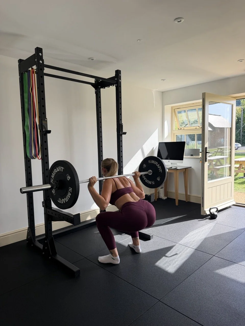 Woman lifting weights in a home gym, squatting with a barbell loaded with weight plates.