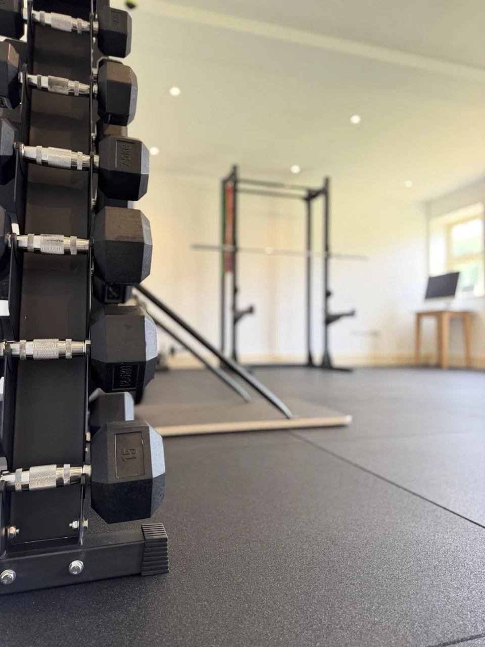 Set of black dumbbells on a shelf in a gym with workout equipment, including a pull-up bar and a laptop on a table, in a well-lit room.
