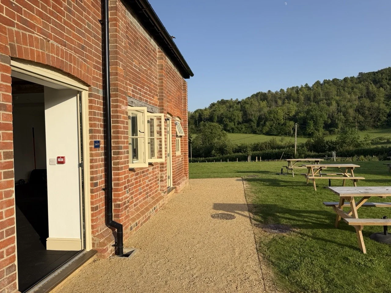 A brick building with an open door and three windows, next to a gravel path, outdoor picnic tables, green grass, and trees with hills in the background under a clear blue sky.