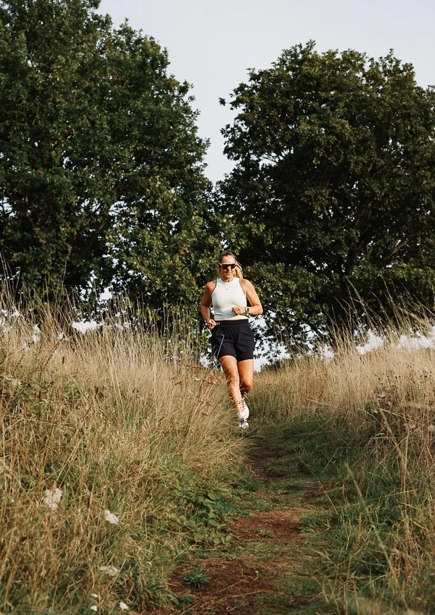 Woman jogging along a dirt trail in a grassy field with tall dry grass and large green trees in the background, wearing sunglasses, a white tank top, black shorts, and running shoes.