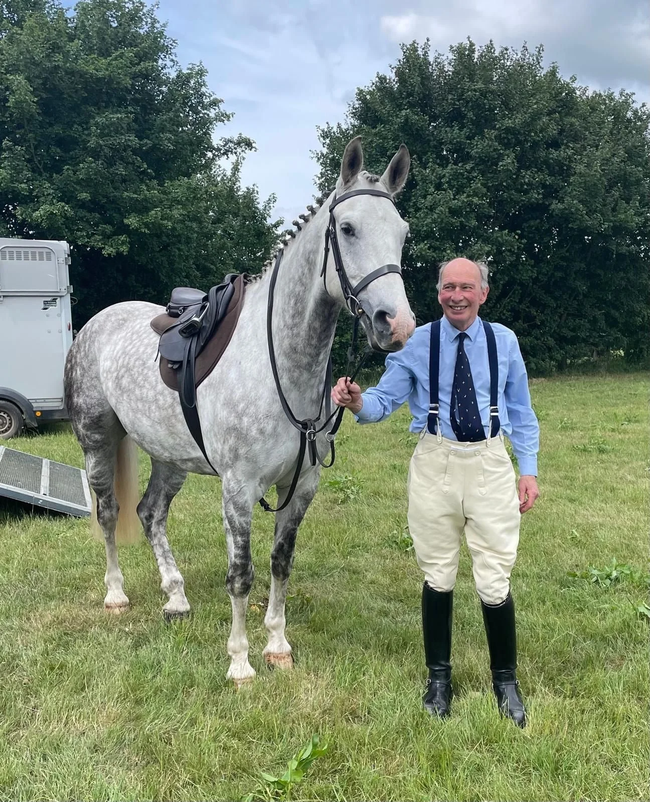 An elderly man in a blue shirt, beige pants, and black riding boots holds a gray horse with a saddle and bridle on a green grassy field. They are standing near trees on a cloudy day.