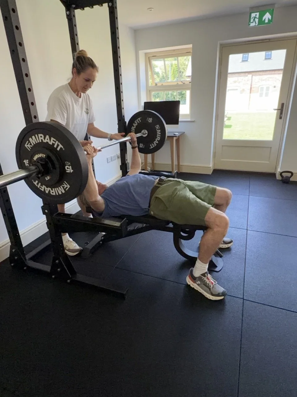 A woman spotter assisting a man doing a bench press in a home gym, with weights on the barbell.