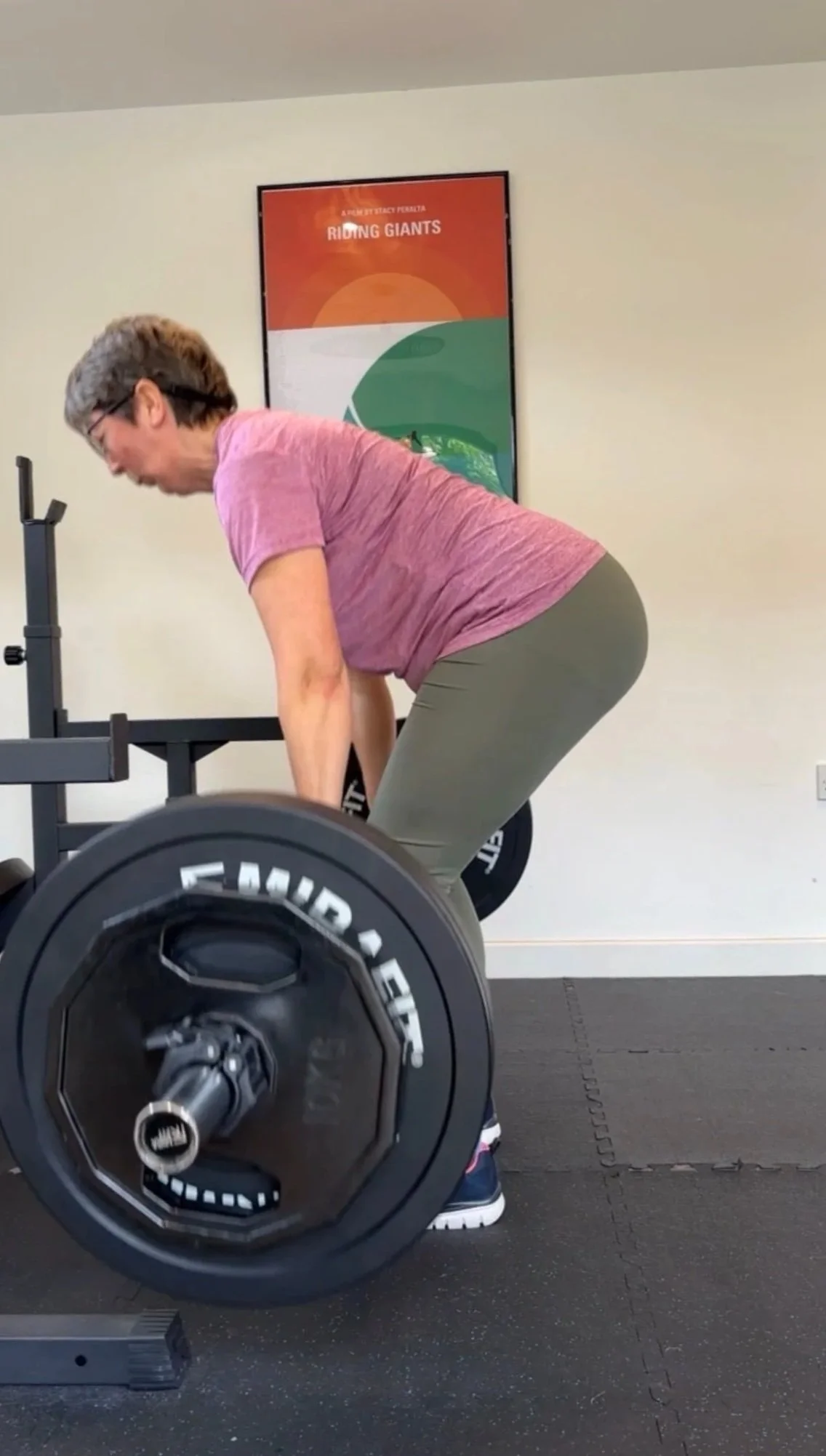 A woman in a pink shirt and gray pants performing deadlifts with a barbell in a gym.