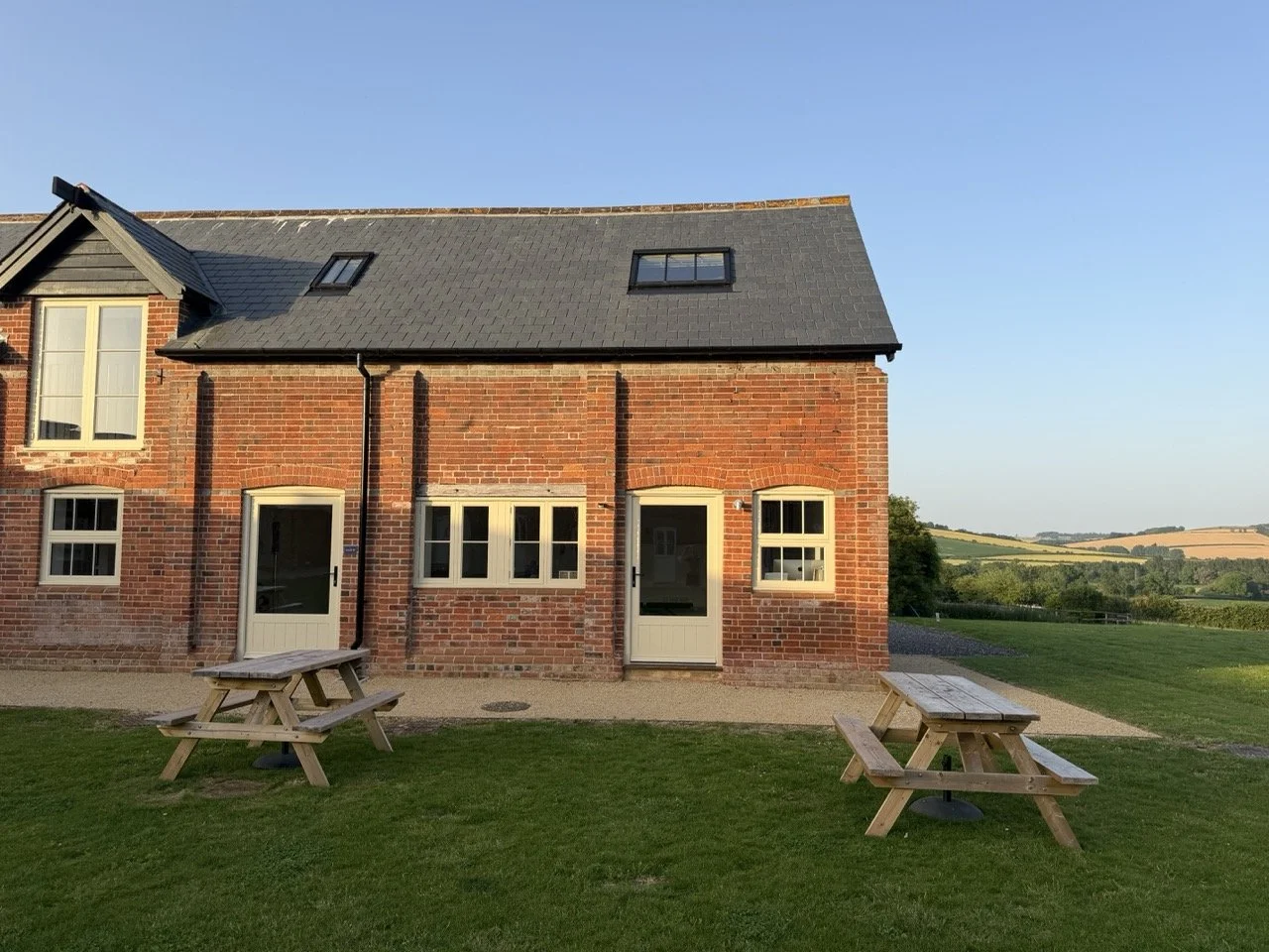 Brick house with black roof, four windows, two doors, and outdoor picnic tables in a grassy yard, in a rural landscape with rolling hills.