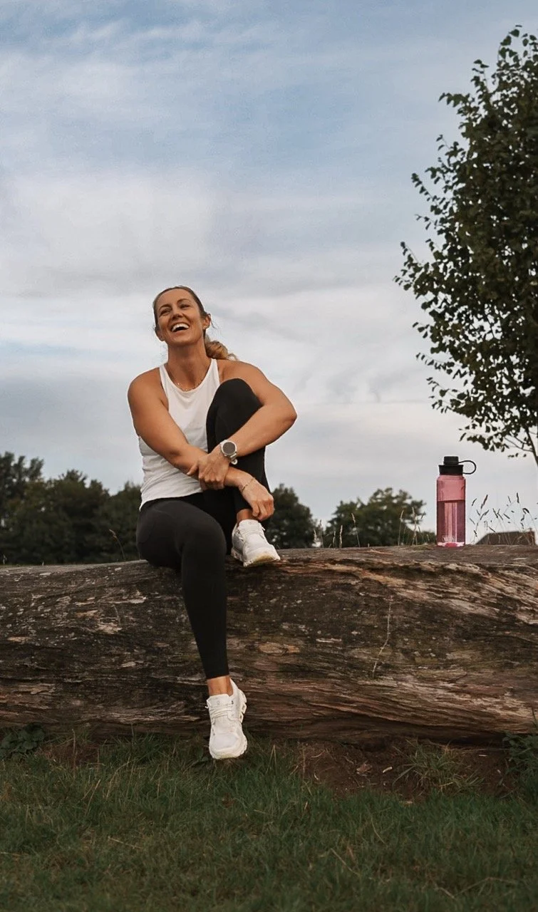 A woman sitting on a fallen log outdoors, smiling and looking up, with a pink water bottle next to her, wearing workout clothes.