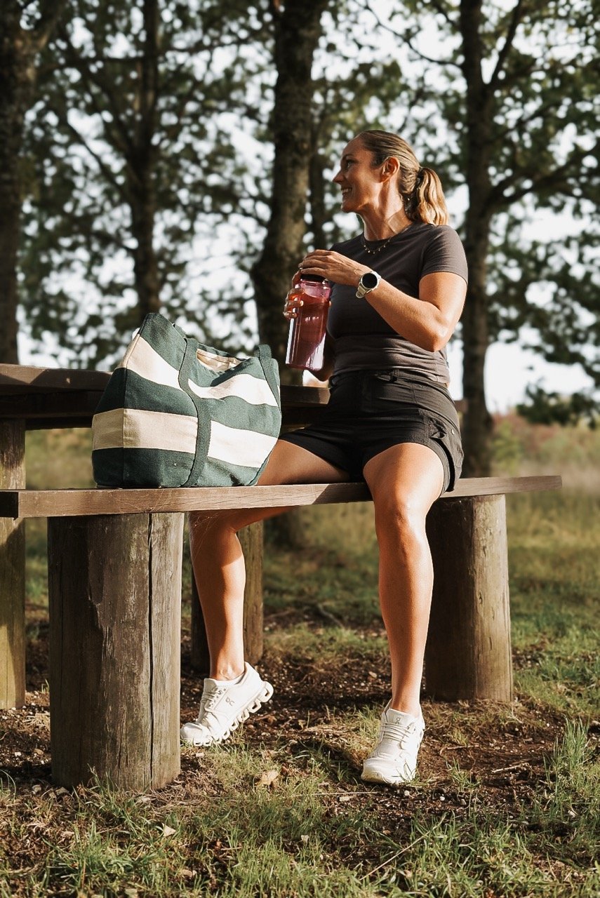 Smiling woman in athletic wear sitting on a wooden park bench, holding a water bottle, with a green and white striped bag beside her, surrounded by trees.