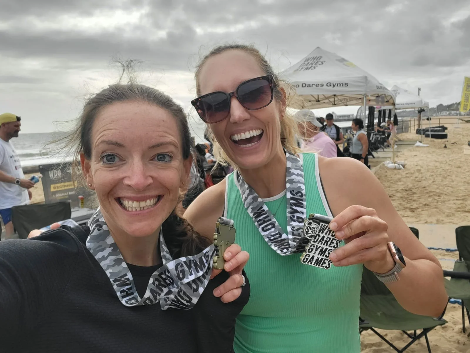 Two women at a beach, smiling and holding medals from a sporting event. The woman on the left has dark hair and a black top. The woman on the right has blonde hair, sunglasses, and a green top. Overcast sky with tents and people in the background.