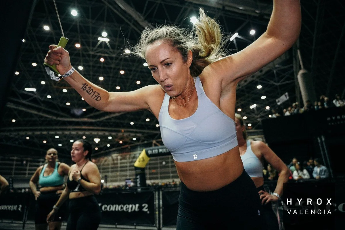 A woman is mid-air, participating in a fitness or CrossFit competition in an indoor arena, with others in workout attire in the background.