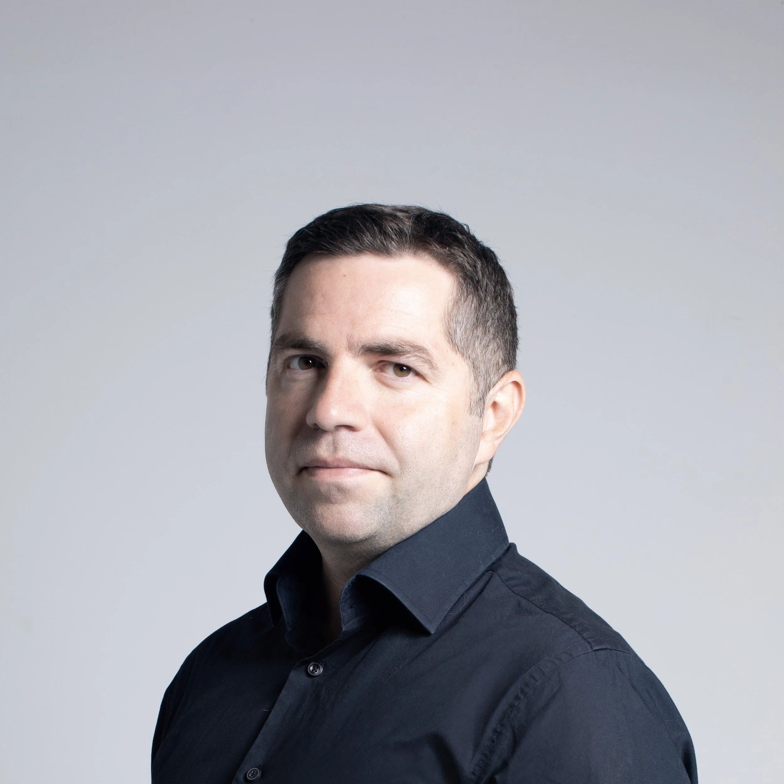 Close-up of a man wearing a black button-up shirt, with short dark hair and light skin, against a plain light gray background.