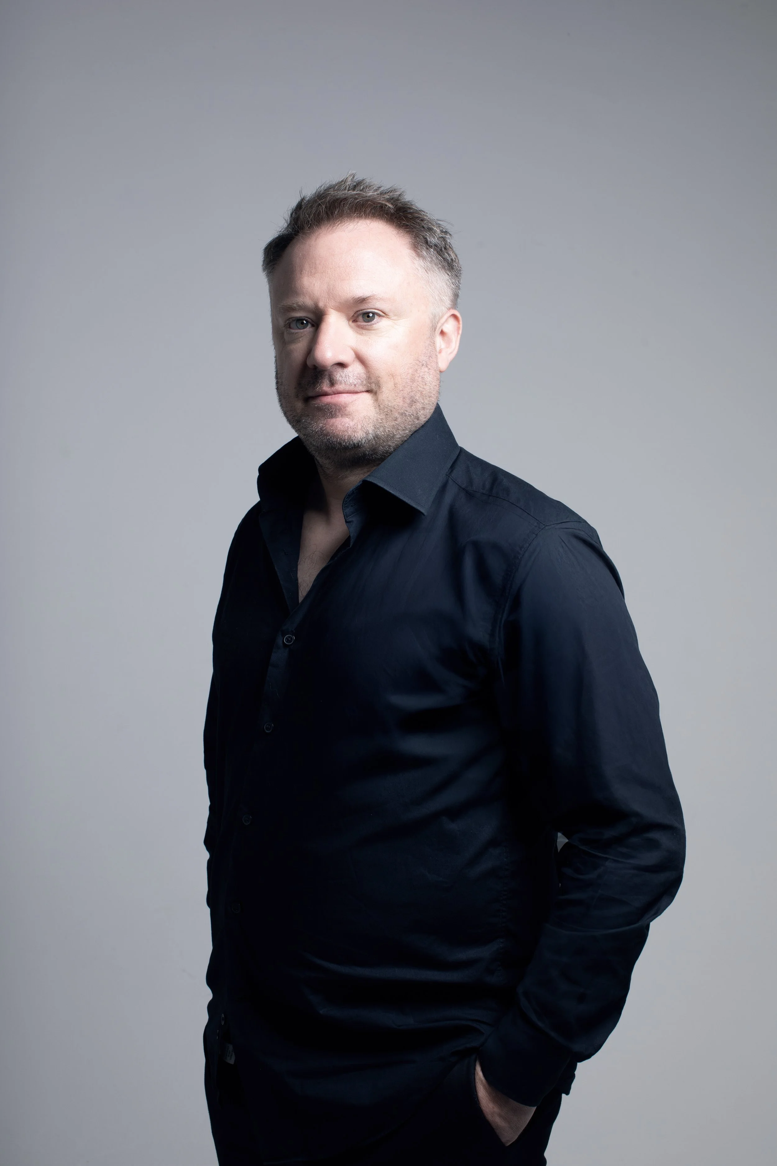 A man with light skin and short, dark hair, wearing a dark shirt, standing against a plain gray background.