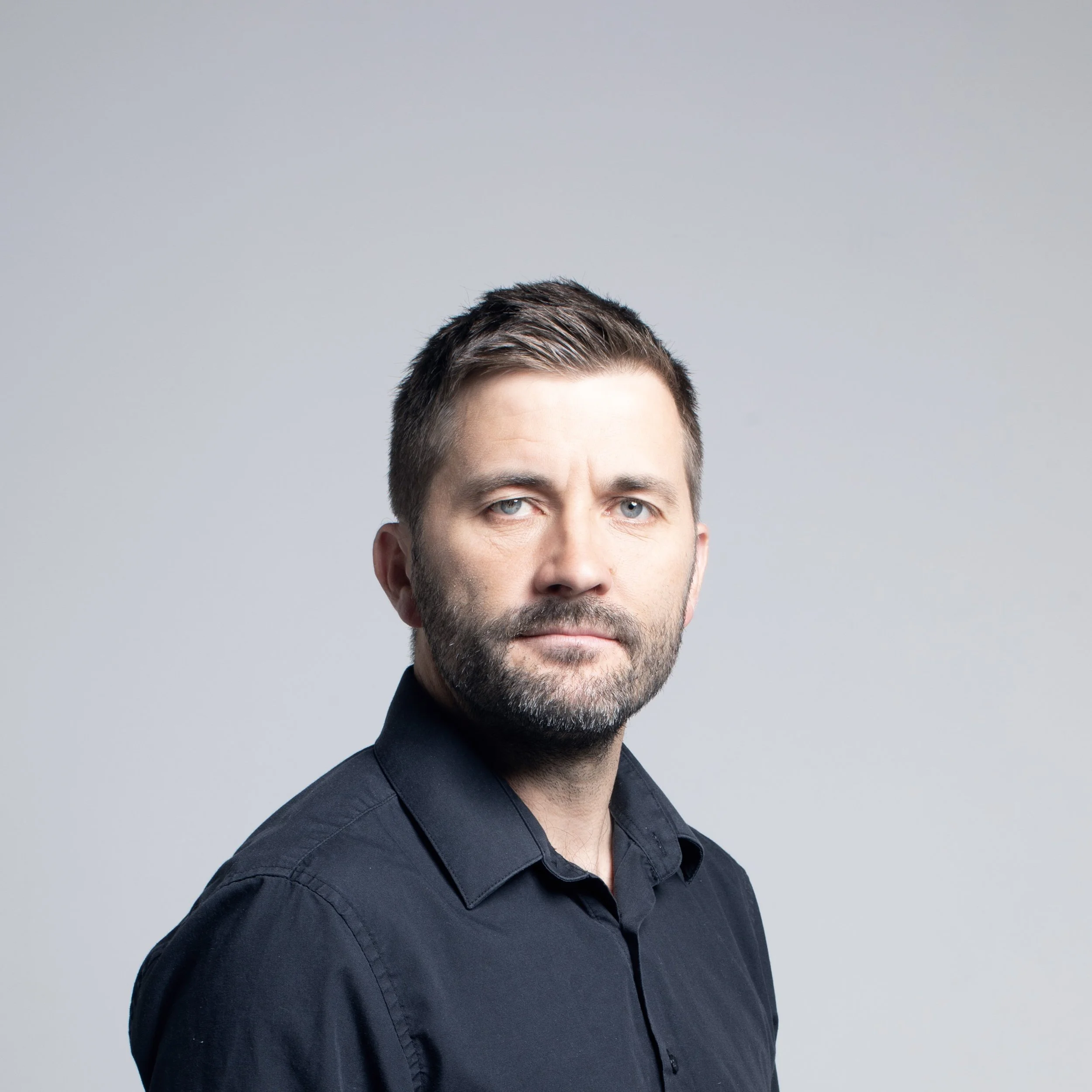 A man with short dark hair, light skin, blue eyes, and a stubbled beard wearing a dark shirt, against a plain gray background.