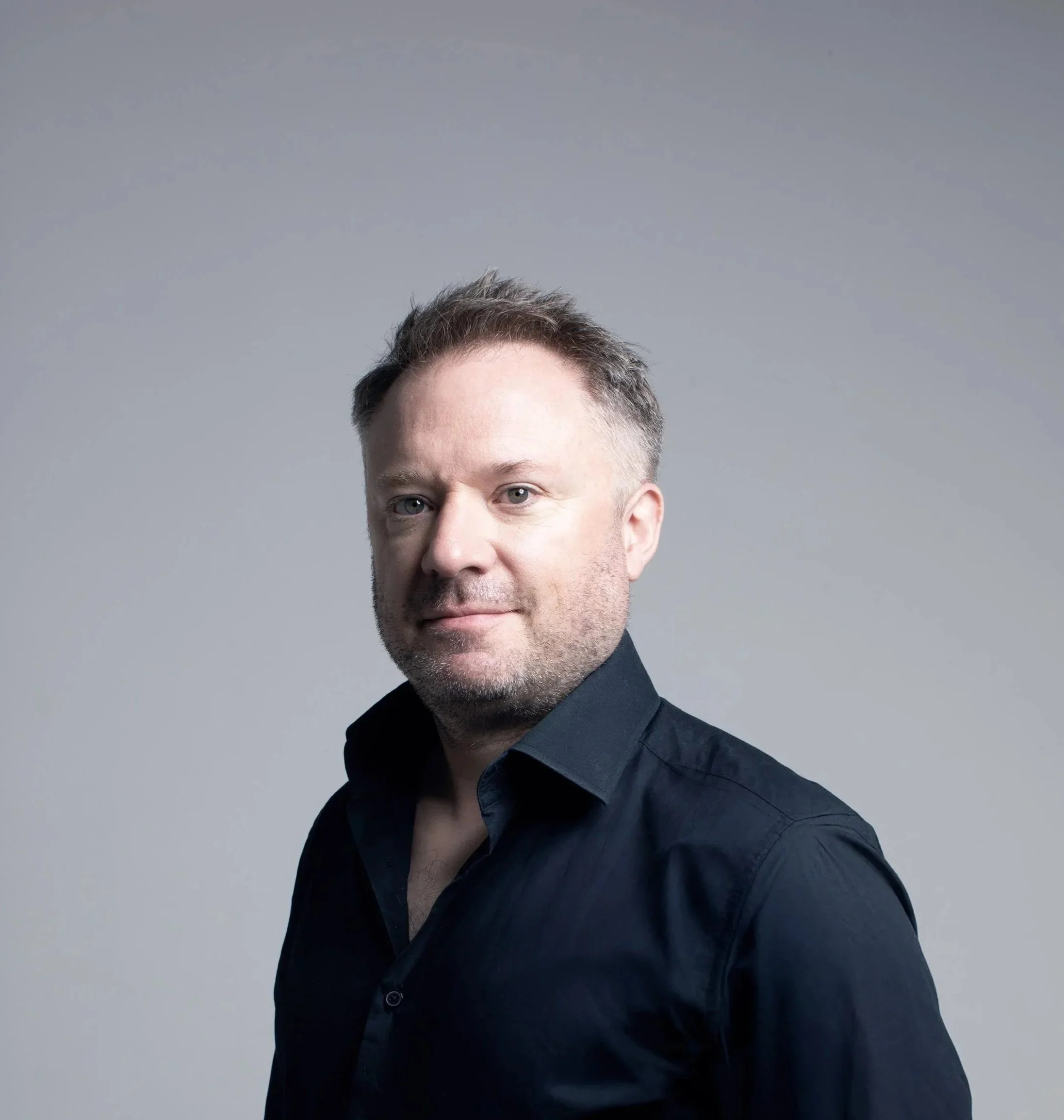 Close-up of a man with short brown hair and blue eyes wearing a dark shirt against a plain gray background.