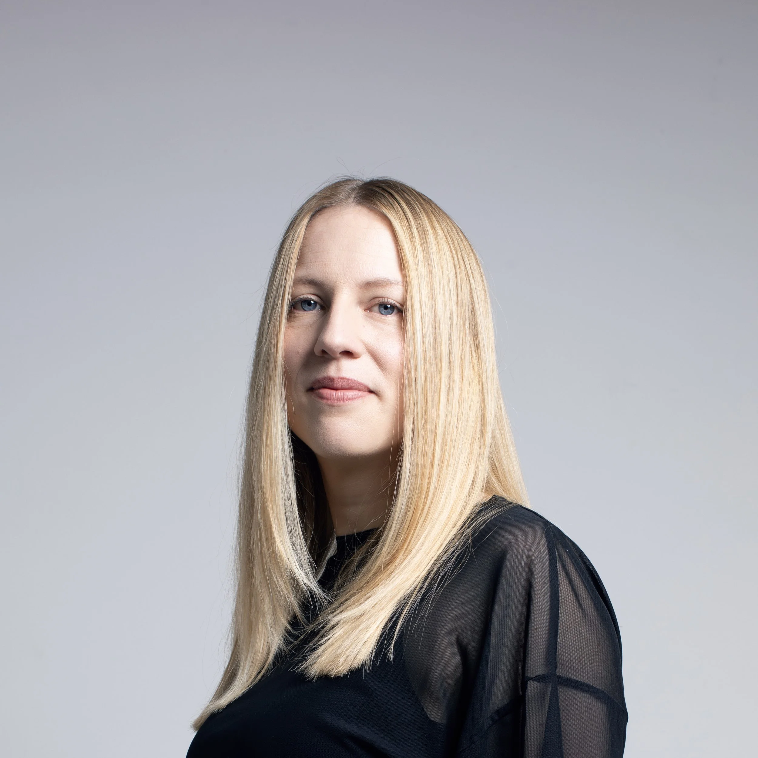Close-up of a woman with long blonde hair and blue eyes, wearing a black top with sheer sleeves, standing against a plain light gray background.