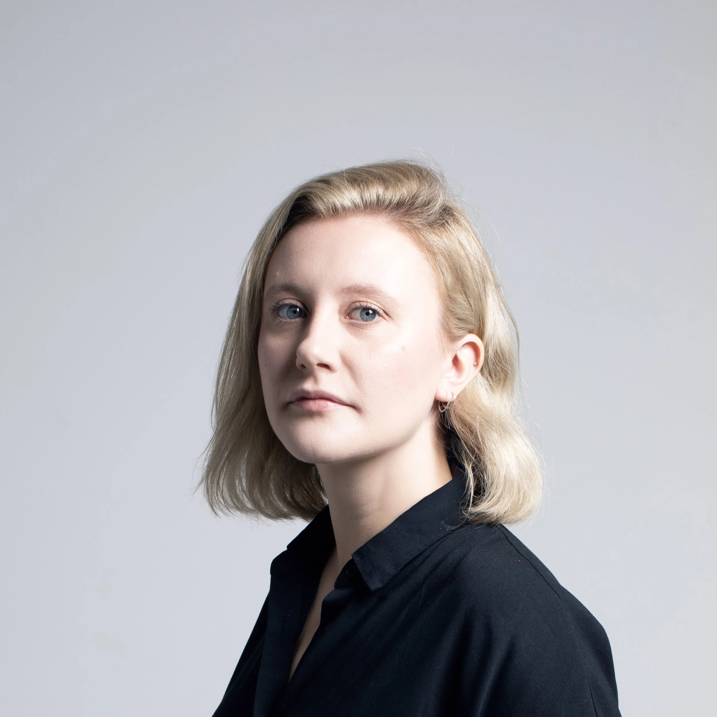Partial portrait of a young woman with blonde hair, blue eyes, and piercings in her ears, wearing a black collared shirt, against a plain light gray background.