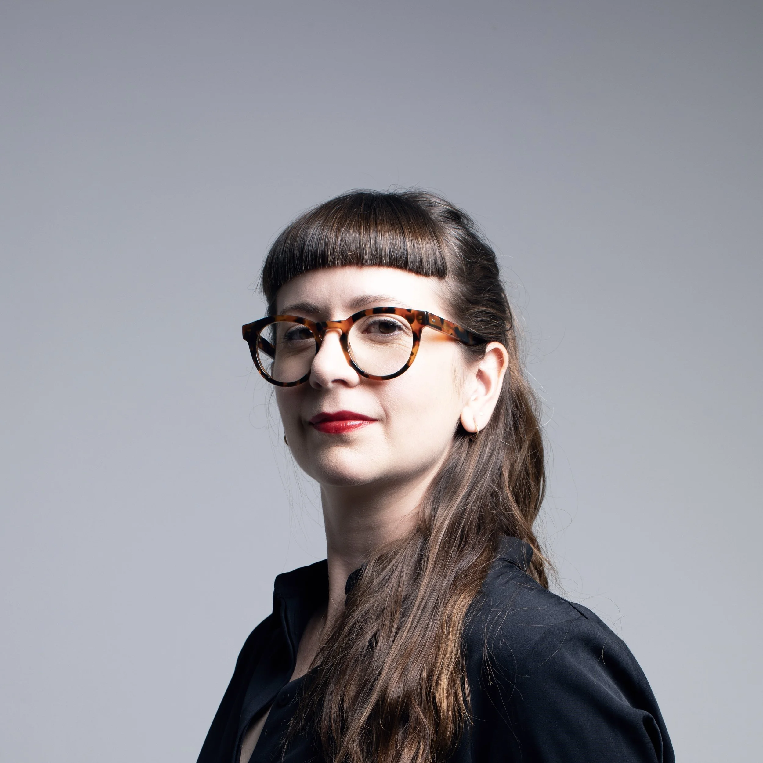 A woman with brown hair, blunt bangs, red lipstick, and tortoiseshell glasses wearing a black blazer against a gray background.