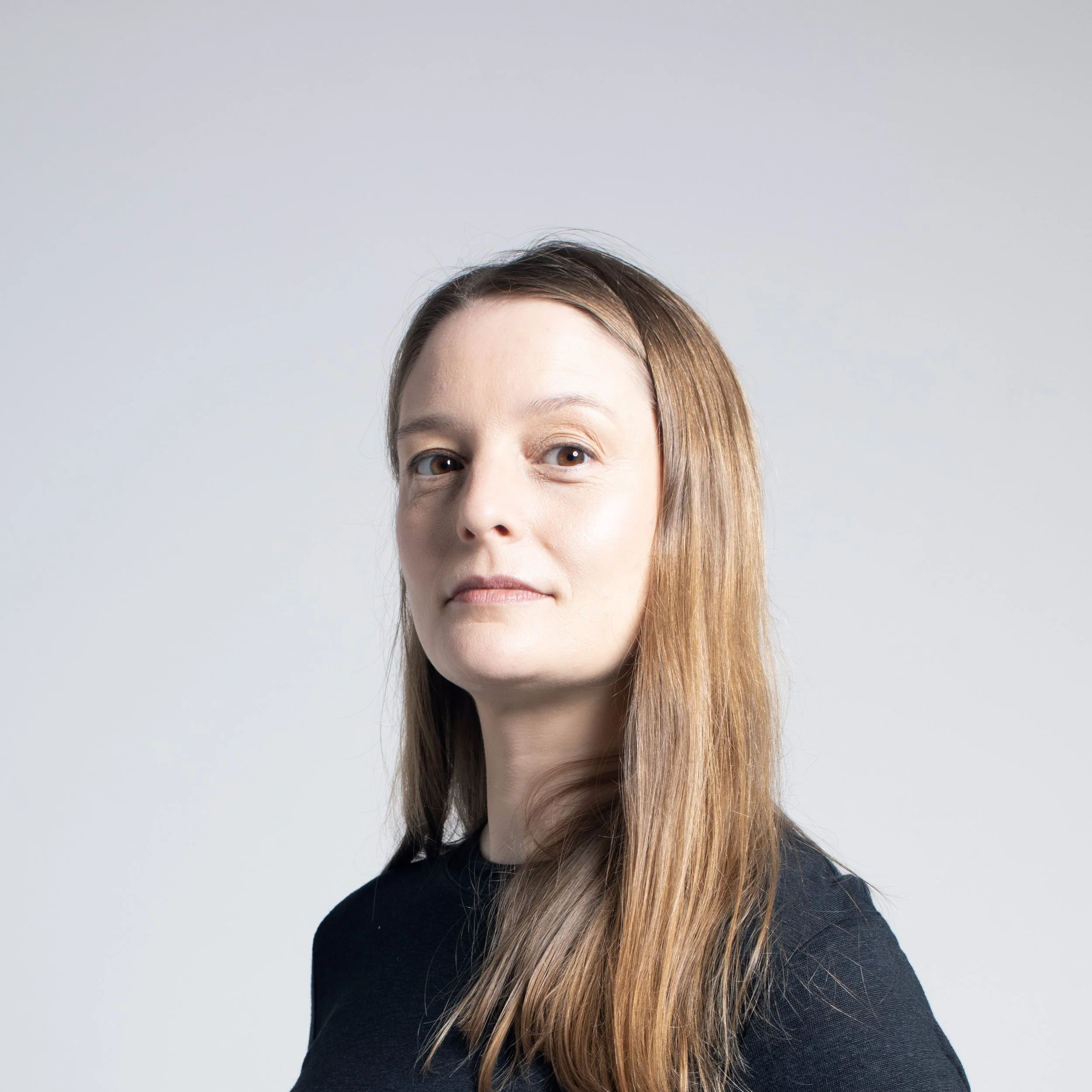 A woman with light skin and long brown hair, wearing a dark blazer, looking directly at the camera, against a plain, light-colored wall.