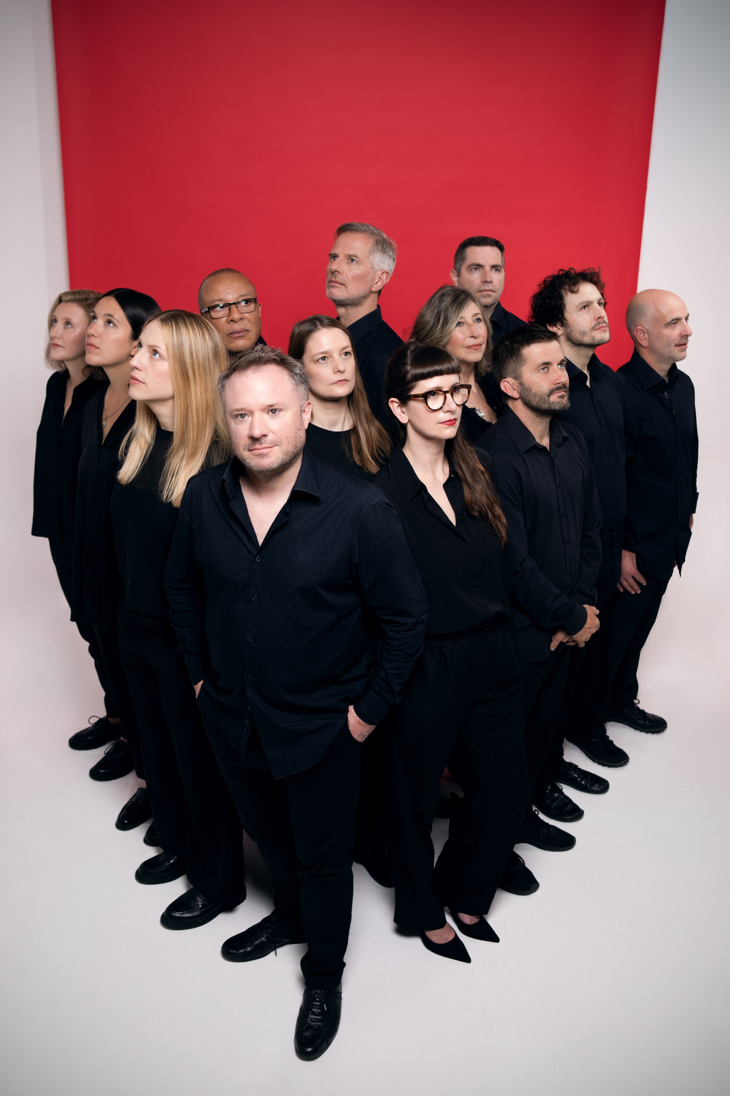 Group of men and women dressed in black clothing, standing in a formation with serious expressions, against a white background with a red wall behind them.