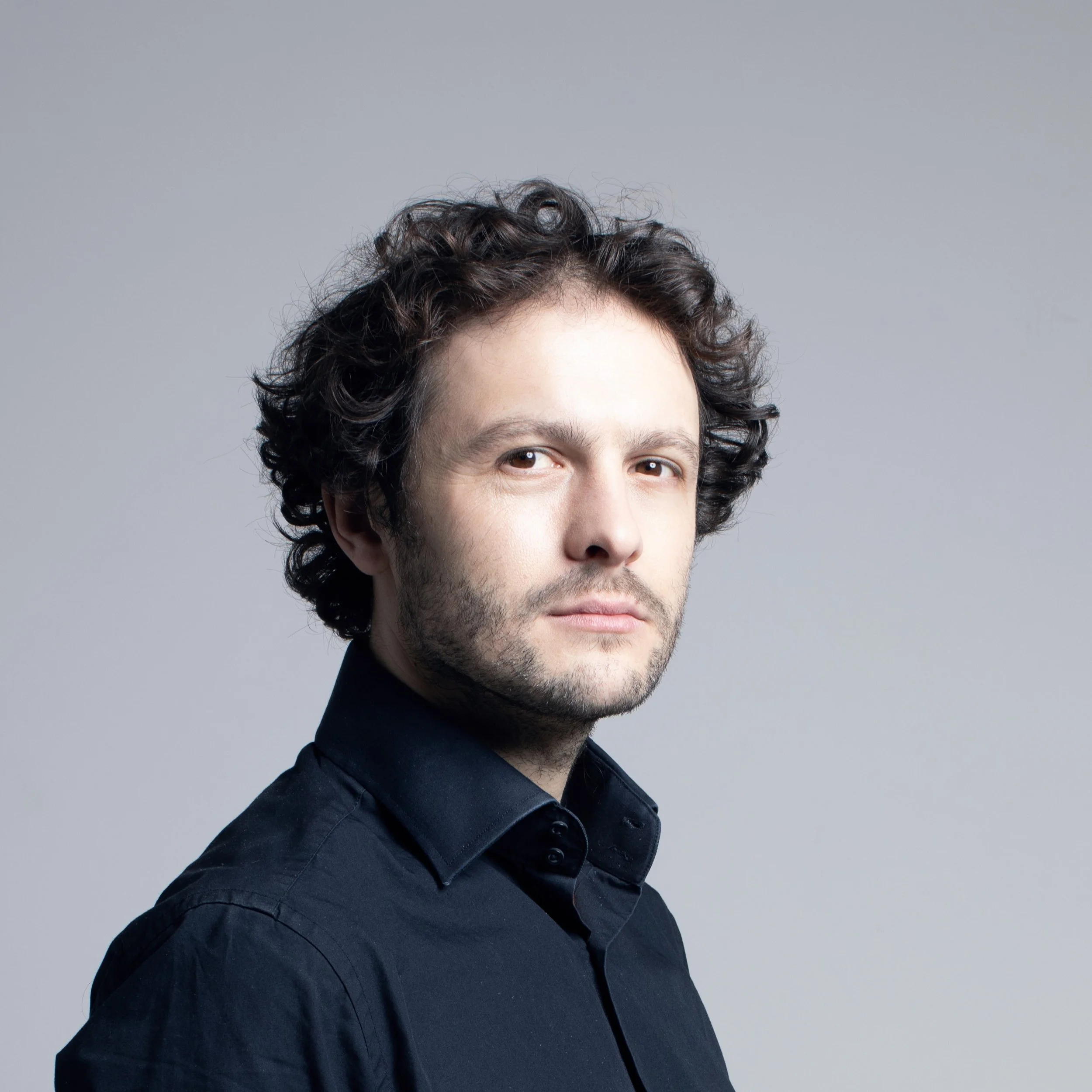 Close-up of a man with dark, curly hair, a short beard, and wearing a black shirt against a gray background.
