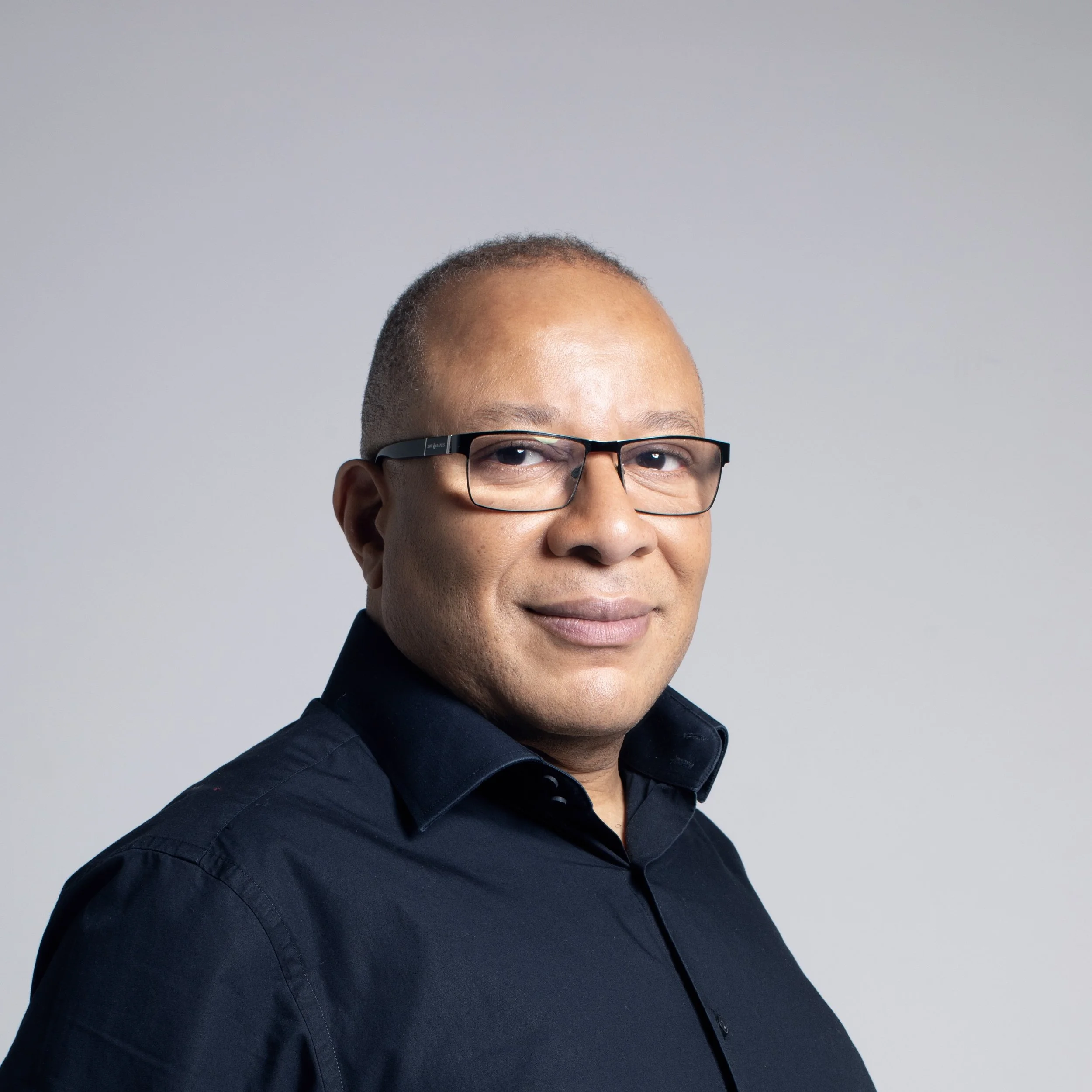 Close-up of a man with short hair, glasses, and a dark shirt, against a plain gray background.