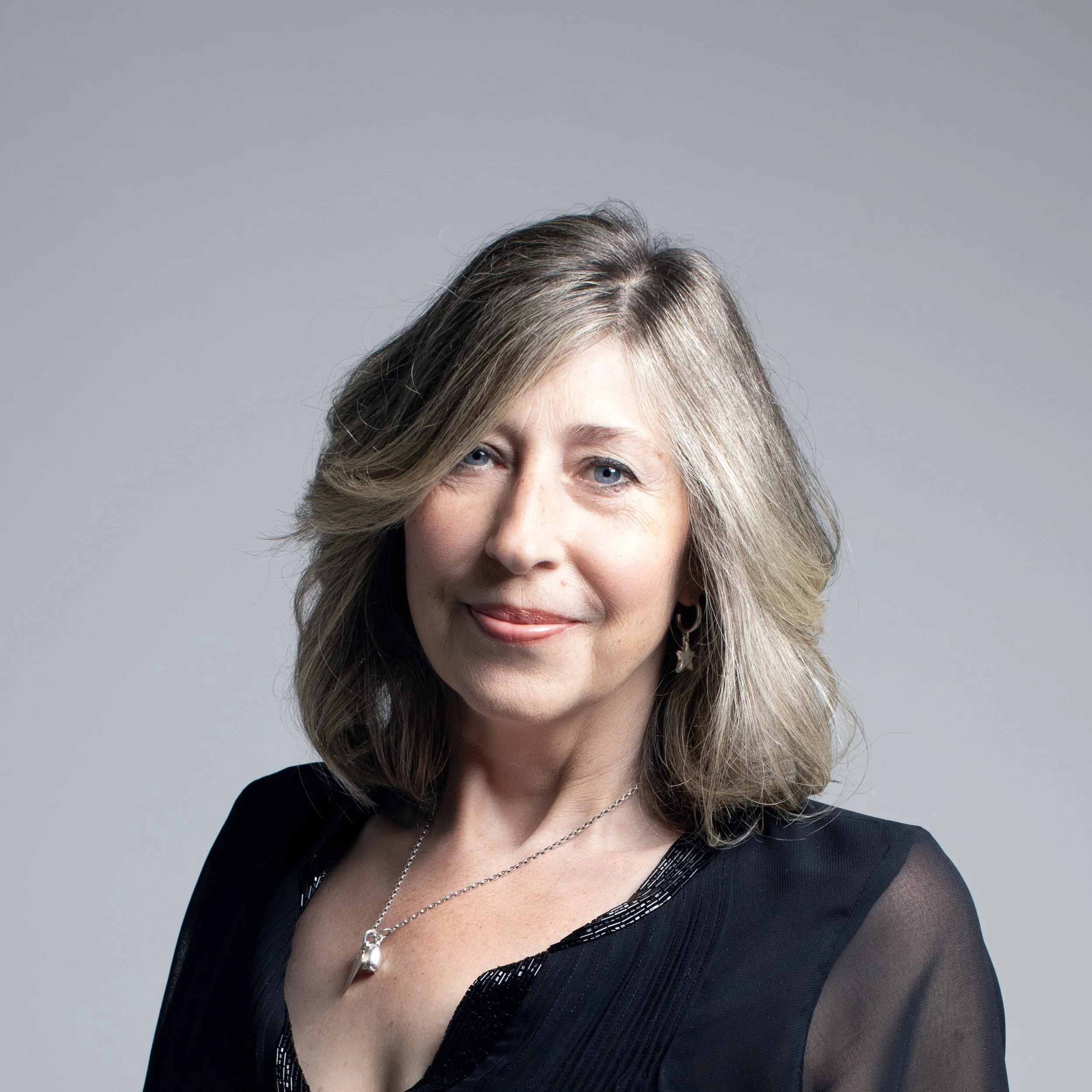 Close-up of a middle-aged woman with grey hair, blue eyes, wearing a black top with decorative beading, and layered silver necklaces, smiling against a plain grey background.