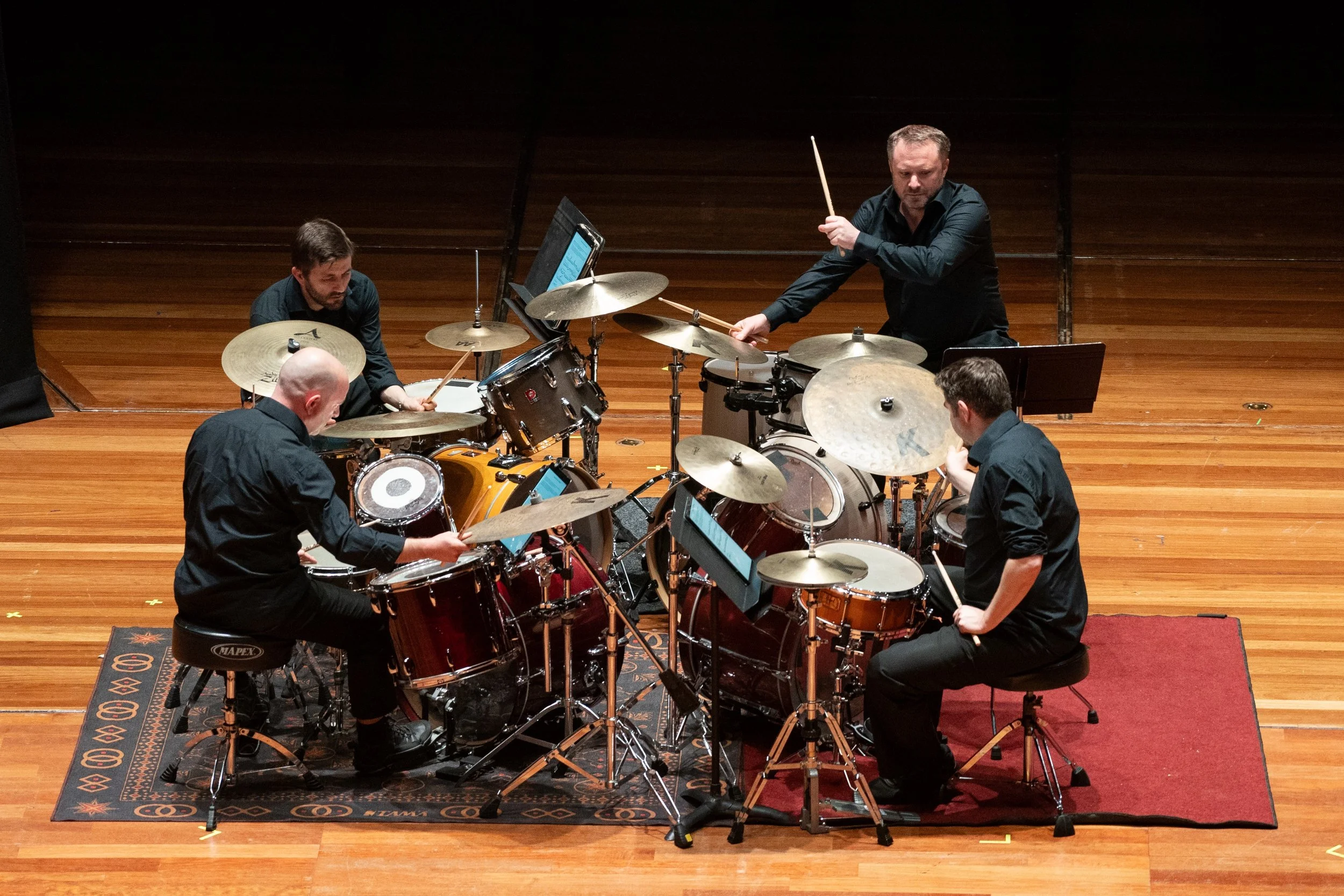 A group of five musicians playing percussion instruments on a wooden stage with music stands and a decorative rug.