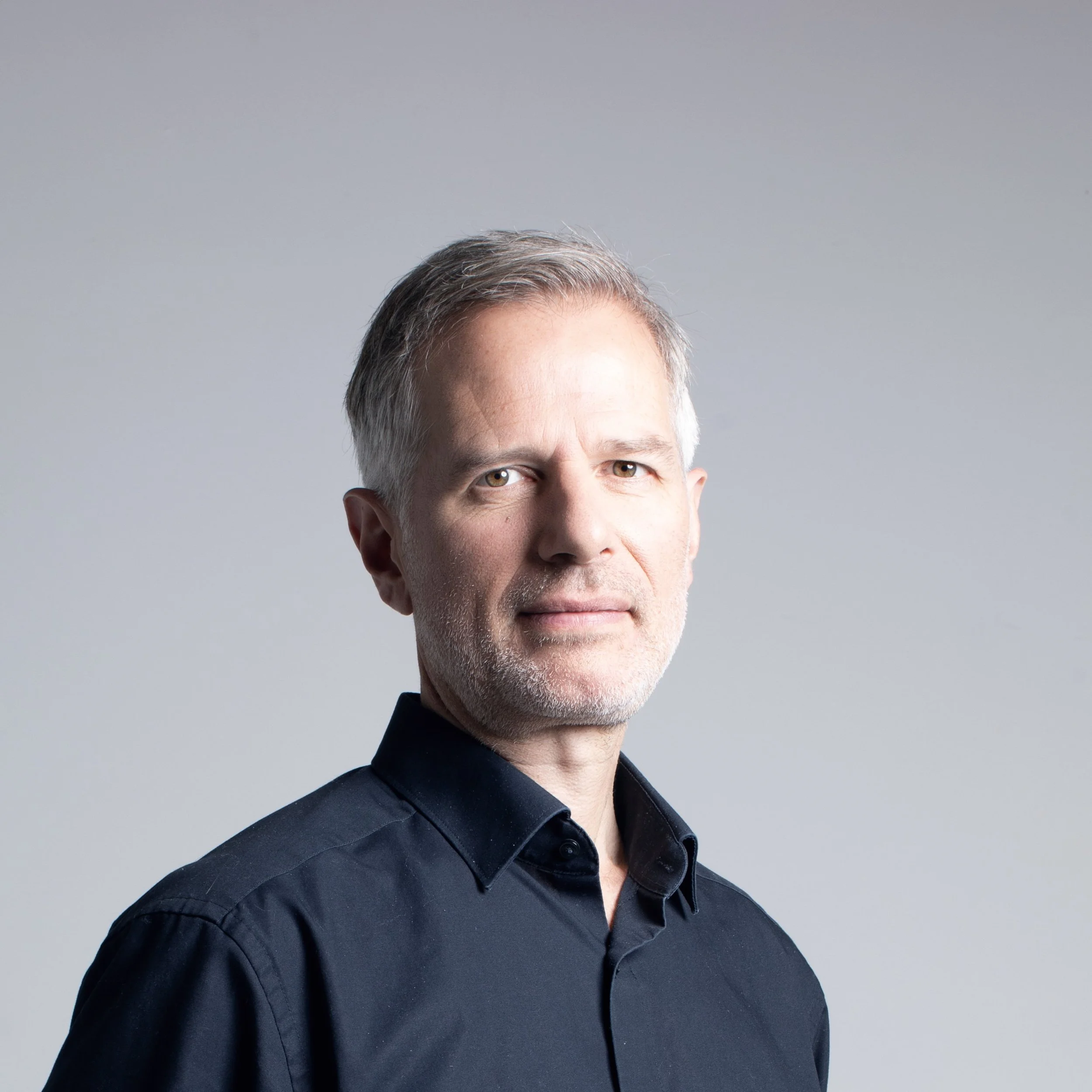 A close-up photo of a man with graying hair wearing a black shirt against a plain light gray background.