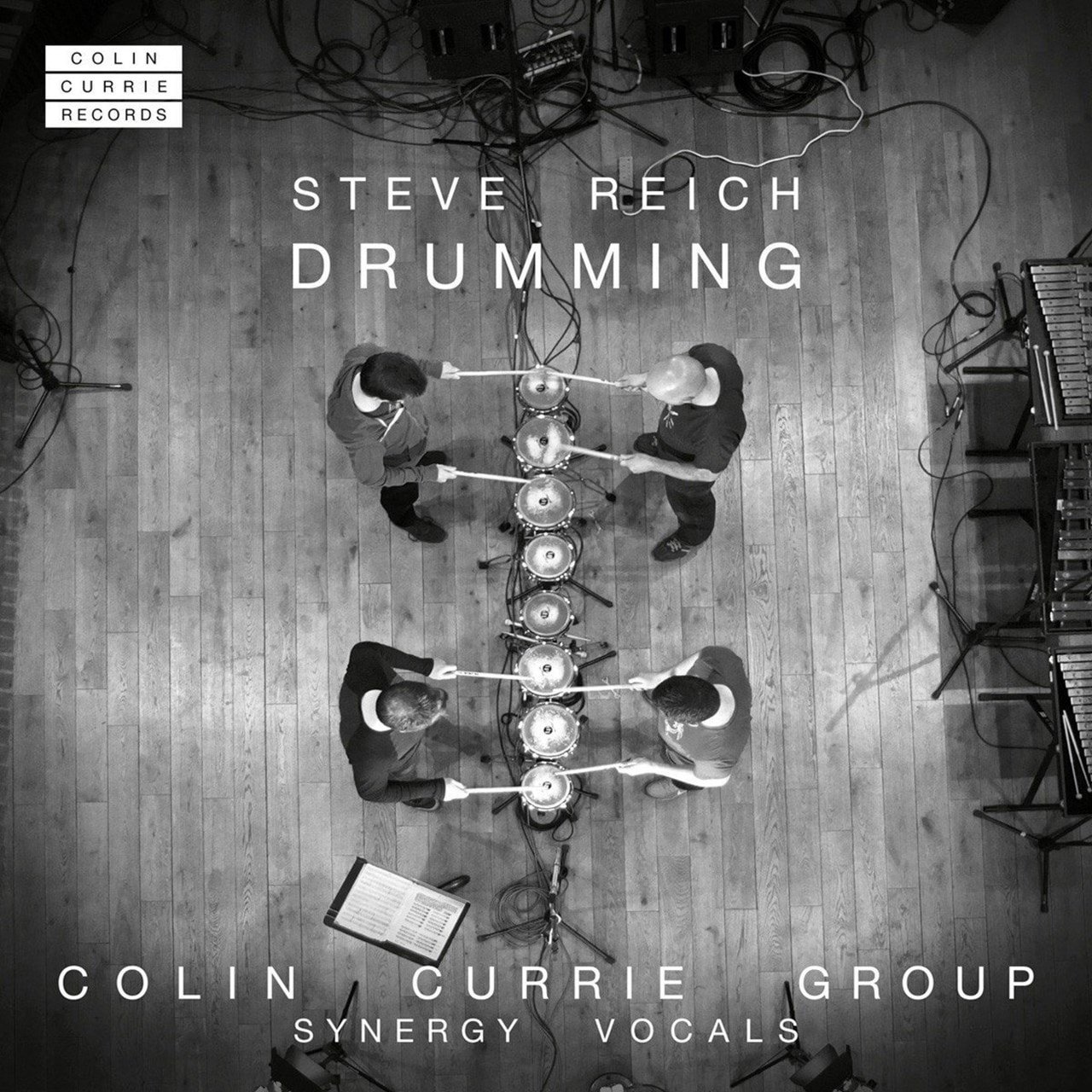 An overhead black-and-white photo shows four people playing tuned drums with drumsticks in a recording studio. There are various cables, microphones, and music equipment around them. On the top left corner, there is a logo for 'Colin Currie Records.' Text on the image reads 'Steve Reich Drumming' and 'Colin Currie Group Synergy Vocals'.