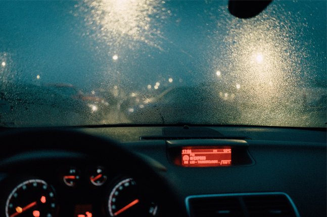 Inside a car during a rainy night, with the windshield covered in raindrops and blurred city lights visible through the windshield.