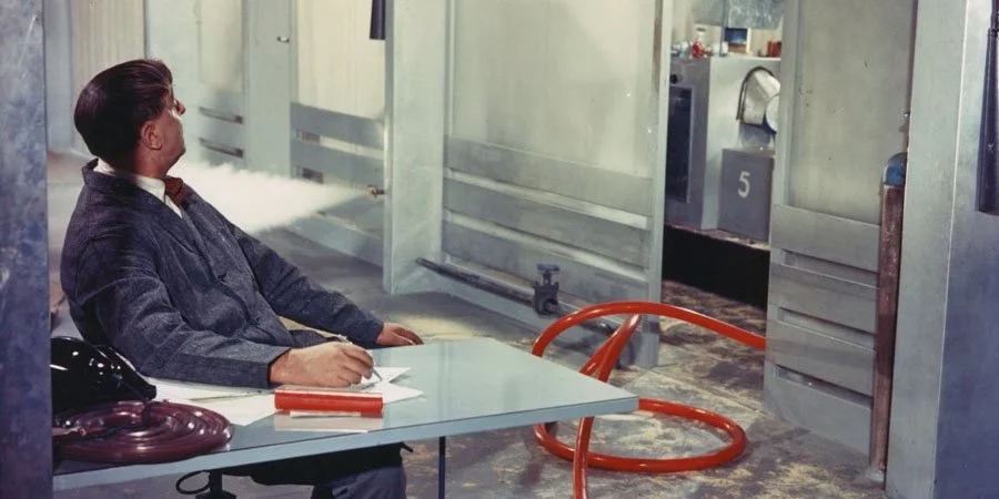 A man in a gray jacket is sitting at a desk with a telephone, papers, and a red book, looking to the left. Behind him, there is a wall with a large door, and in the background are industrial or underground-like surroundings with pipes and metal structures.