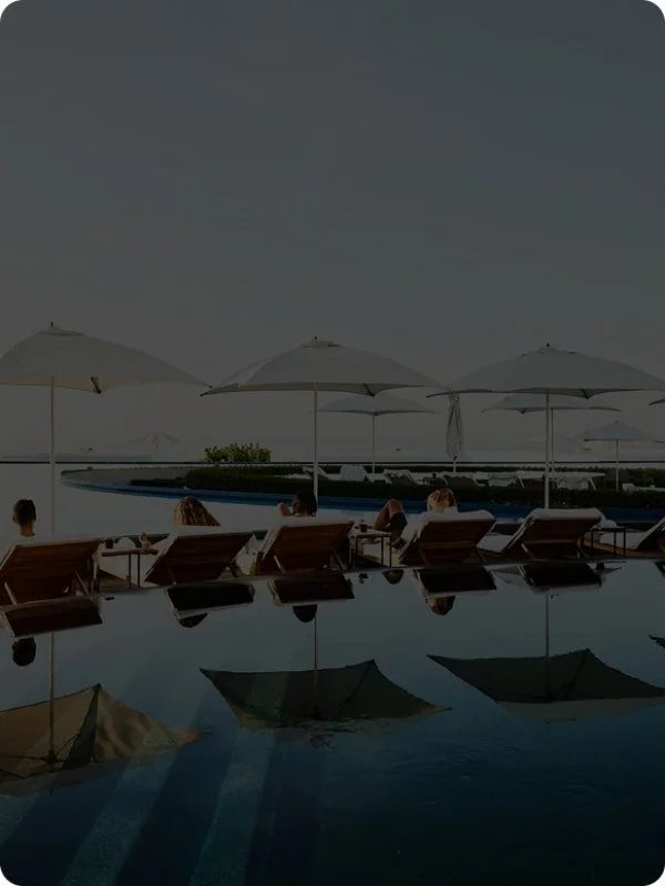 People lounging on chairs under umbrellas poolside at a resort or hotel.