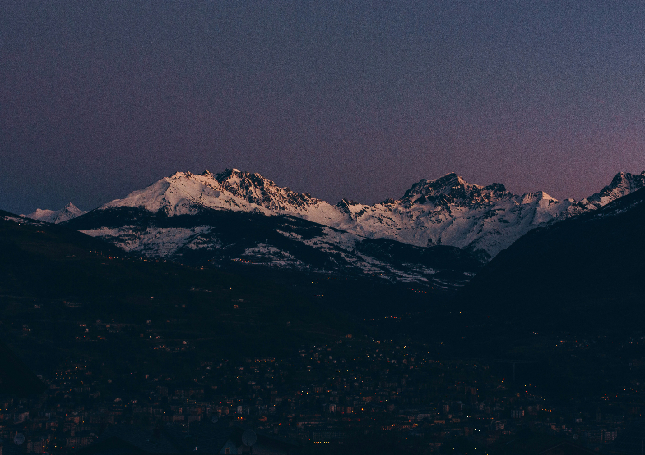 Nighttime view of snow-capped mountains under a dark sky with some city lights visible at the base.
