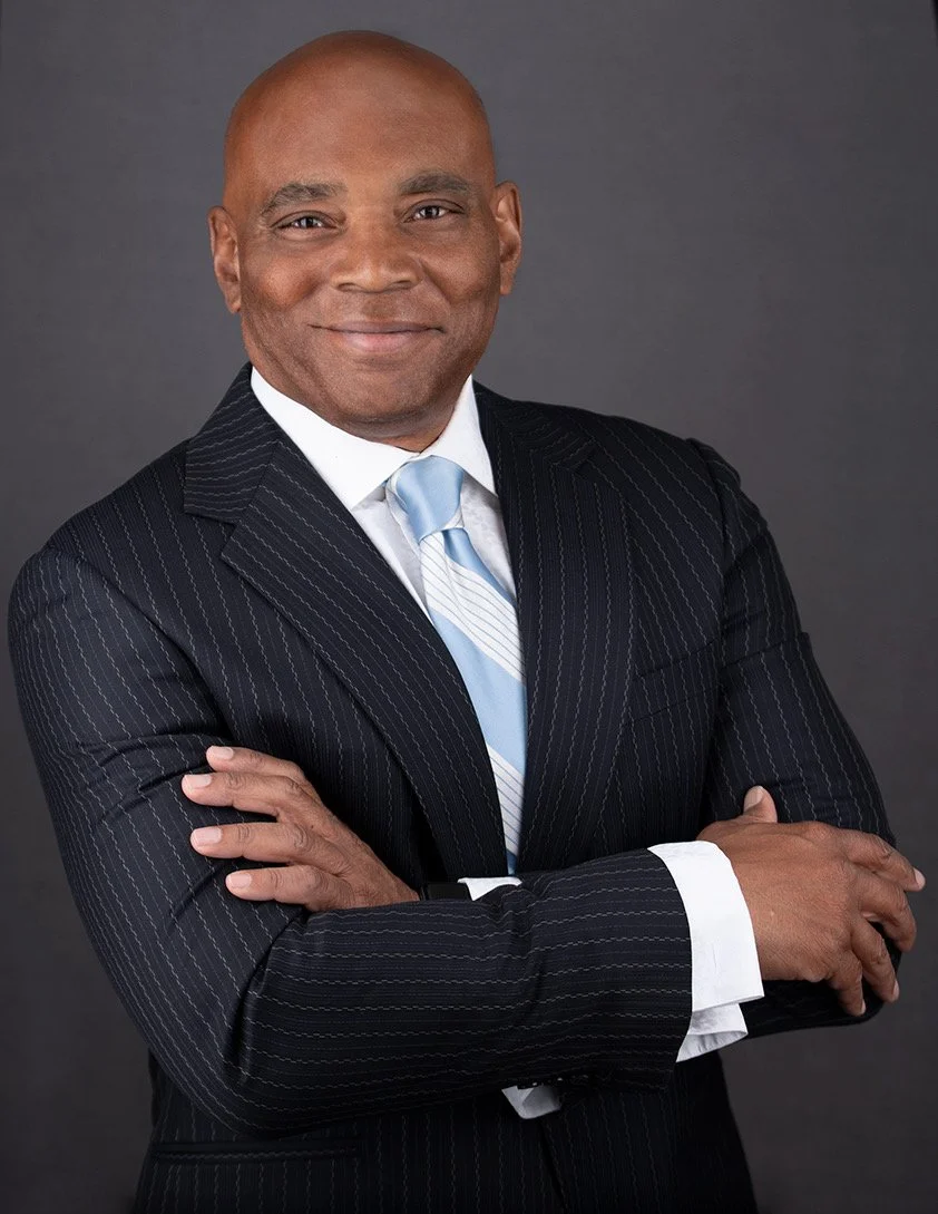 A middle-aged African American man wearing a navy blue pinstriped suit, white shirt, and light blue tie, standing with crossed arms and smiling, against a dark gray background.